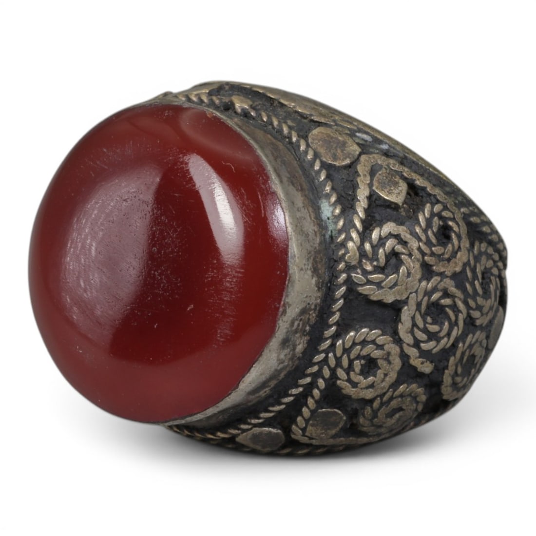 Antique Central Asian Silver and Carnelian Tribal Ring, 19th-Early 20th Century, 60AJ: The ring is set with a large, smooth cabochon carnelian stone, secured within a silver mount. The mount is decorated with pseudo-filigree wirework and applied coiled motifs, set against a darkened bac