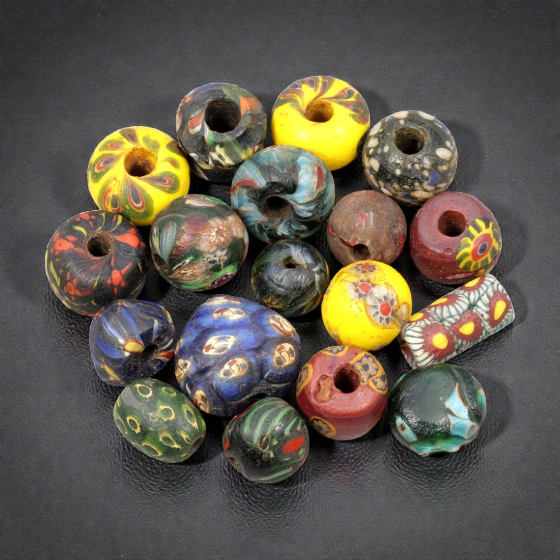 A Lot of 17 Antique/ Vintage Artisan Glass Trade Beads, 26WM: A curated collection of 17 glass trade beads, varying in form from spherical, barrel, and cylindrical to polygonal, all with central perforations. The beads feature intricate mosaic, trailed, and appl