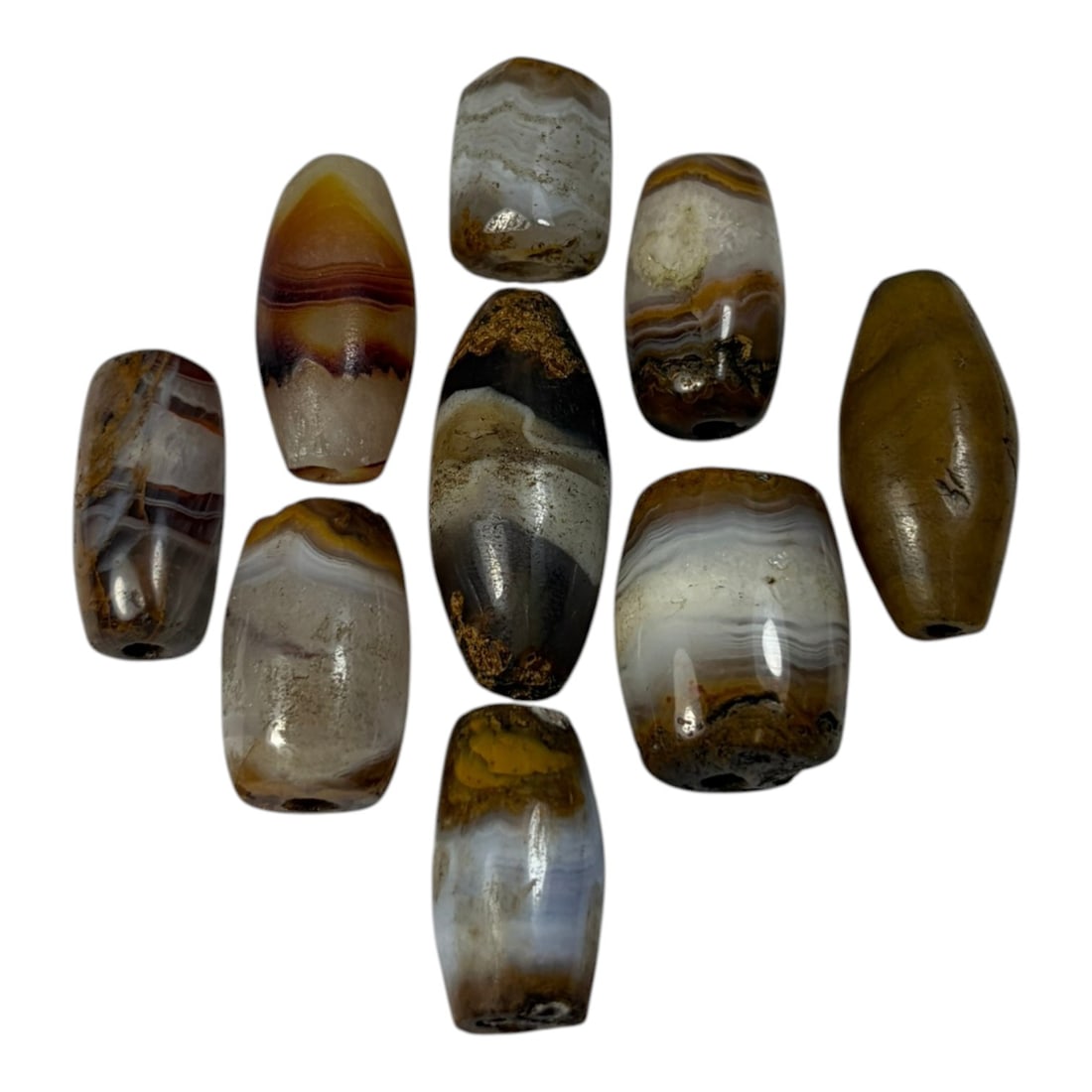 A Lot of 9 Himalayan Banded Agate Beads, Ancient to Antique Period, 45AN: Comprising lot of barrel and ovoid beads, each carved from banded agate with natural tonal variations, many displaying characteristic concentric, linear, and dendritic patterns. The surfaces exhibit s
