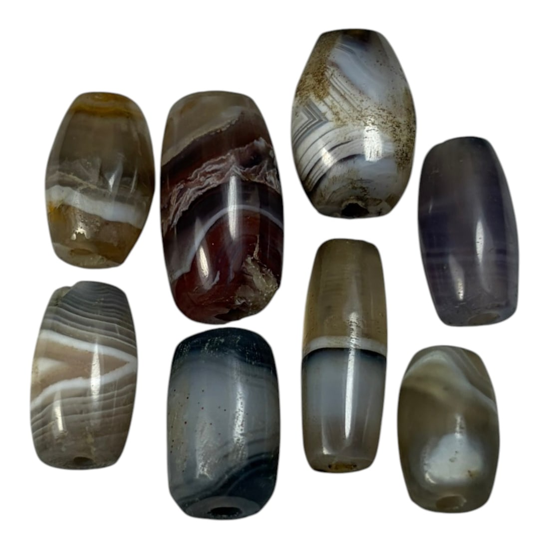 A Lot of 9 Himalayan Banded Agate Beads, Ancient to Antique Period, 54AN: Comprising lot of barrel and ovoid beads, each carved from banded agate with natural tonal variations, many displaying characteristic concentric, linear, and dendritic patterns. The surfaces exhibit s