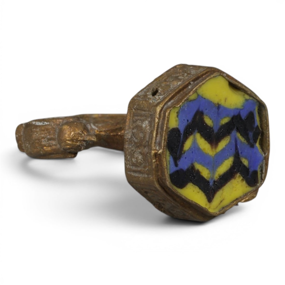 Antique Qajar Dynasty Gilt Glass Ring, US Size 6, 5AJ: A gilt-metal ring of the Qajar period, US ring size 6, featuring a hexagonal glass inlay with a vibrant yellow, blue, and black zigzag pattern. The bezel is adorned with embossed geometric motifs, whi