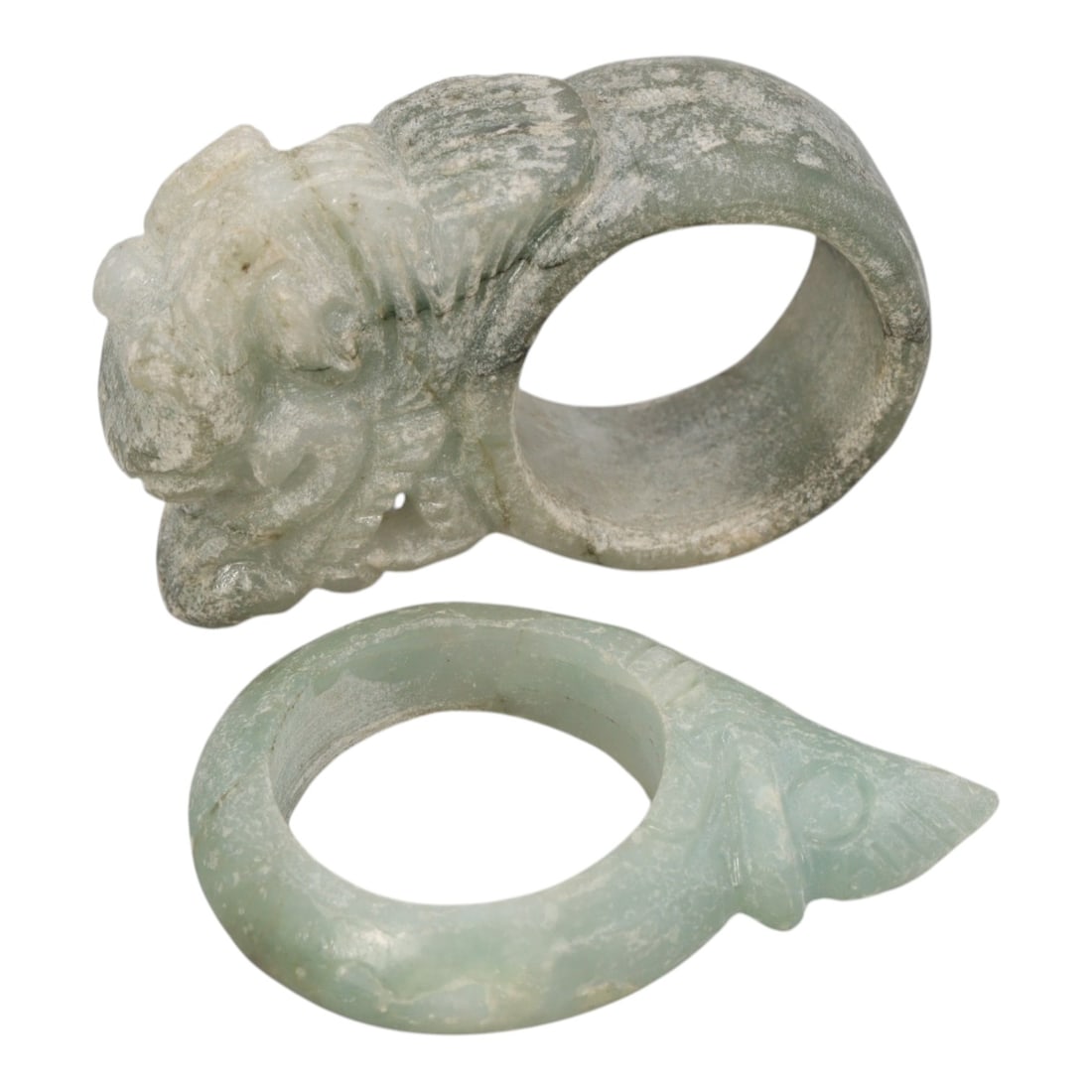A Pair of Type A Jadeite Rings - Carved Animal, 16WS: This lot includes two type A jadeite rings, both carved from pale celadon-green jadeite with natural surface patina and mineral inclusions. The first ring features a detailed mythical guardian beast h