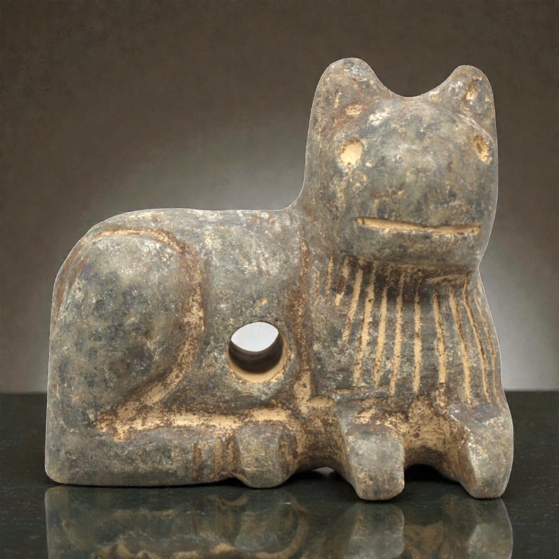 Ancient Near Eastern Stone Feline Amulet, Circa 3rd-2nd Millennium BCE, 89ZP: Carved from grey stone with natural inclusions and earthy patination, the amulet depicts a recumbent feline figure. The form is stylized with simplified facial features, including incised eyes and a l