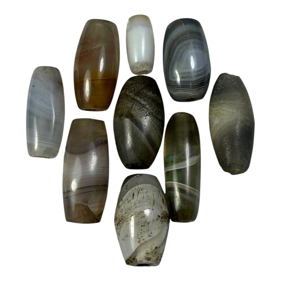 A Lot of 9 Himalayan Banded Agate Beads, Ancient to Antique Period, 50AN: Comprising lot of barrel and ovoid beads, each carved from banded agate with natural tonal variations, many displaying characteristic concentric, linear, and dendritic patterns. The surfaces exhibit s