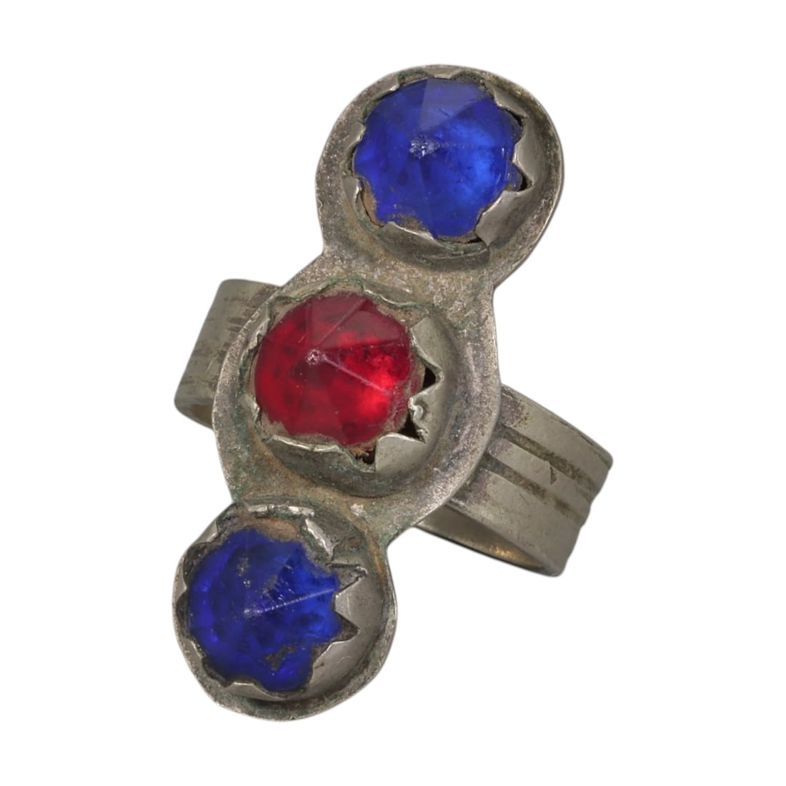 A Berber Silver Three-Stone Ring, Late 19th-Early 20th Century, 38AK: Crafted from silver, featuring a wide band with parallel incised linear detailing. The vertical bezel holds three faceted stones: two blue and one red, set in scalloped bezels. The metal surface shows