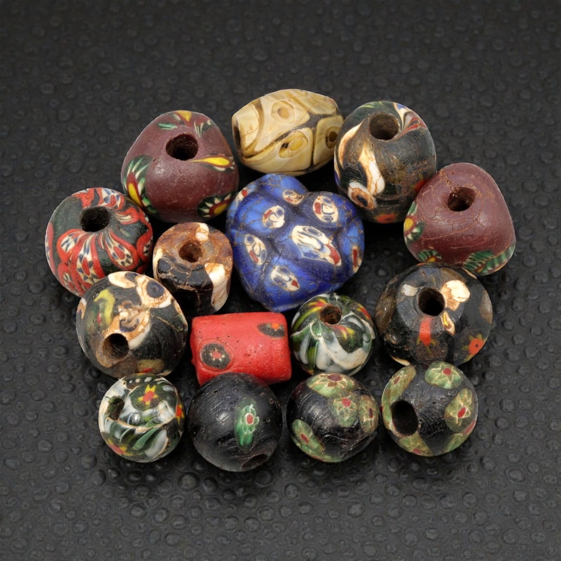 A Lot of 15 Antique/ Vintage Artisan Glass Trade Beads, 28WM: A curated collection of 15 glass trade beads, varying in form from spherical, barrel, and heart-shaped to polygonal, all with central perforations. The beads feature intricate mosaic, trailed, and app