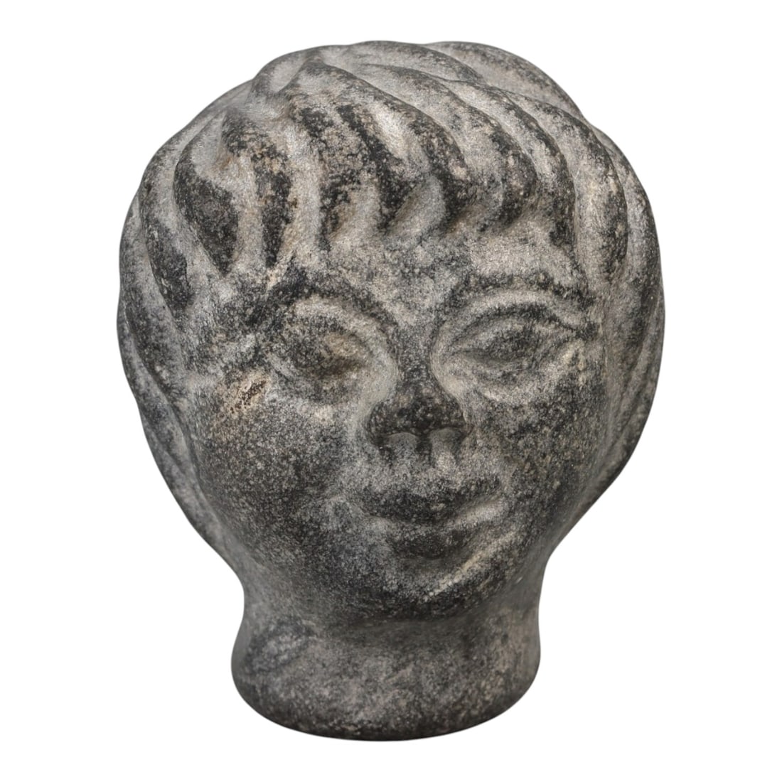 A Roman Stone Head, Circa 2nd-3rd Century CE, 3FT: Carved from a granular, dark grey stone with natural light inclusions, the object is a complete head, not a fragment, depicting a youthful male figure. The face is rendered with stylized features: alm