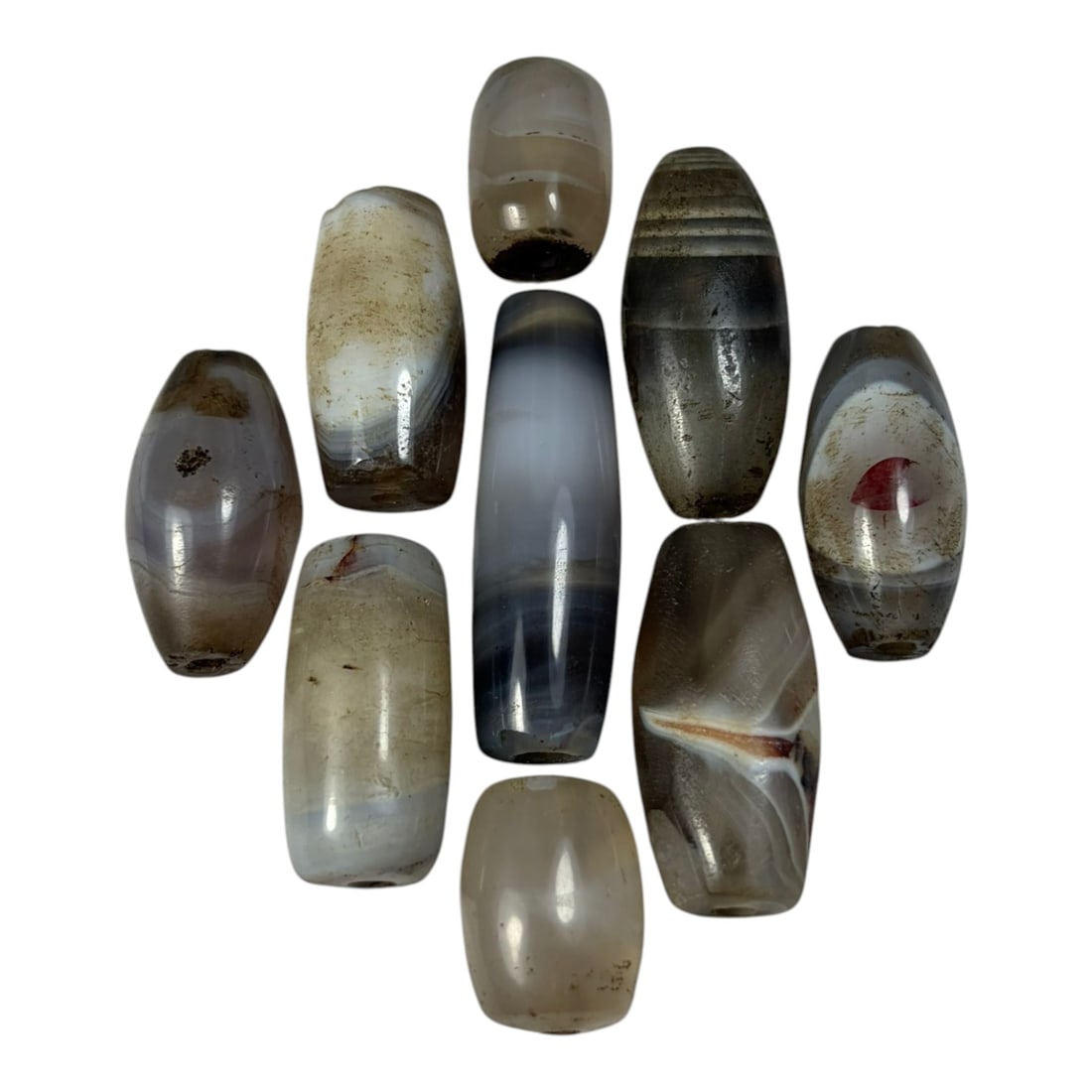 A Lot of 9 Himalayan Banded Agate Beads, Ancient to Antique Period, 40AN: Comprising lot of barrel and ovoid beads, each carved from banded agate with natural tonal variations, many displaying characteristic concentric, linear, and dendritic patterns. The surfaces exhibit s