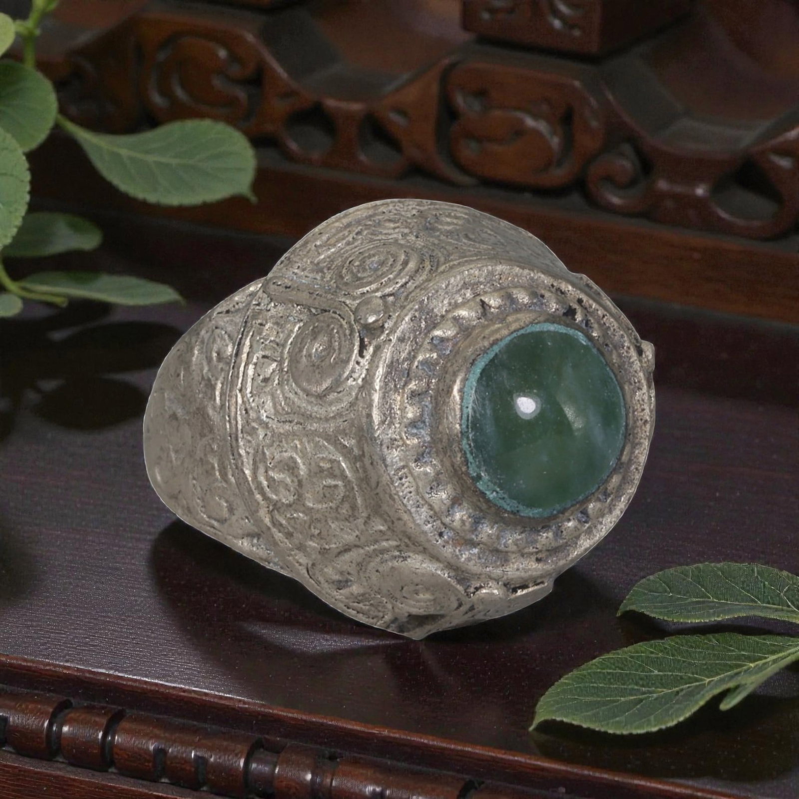 A Middle Eastern Silver Plated Green Agate Ring, Late 19th-Early 20th Century, 18AK: The ring is centered with a round cabochon green agate stone set within a domed bezel featuring a raised beaded border. The bezel and shoulders are engraved with stylized spiral and foliate motifs. Th