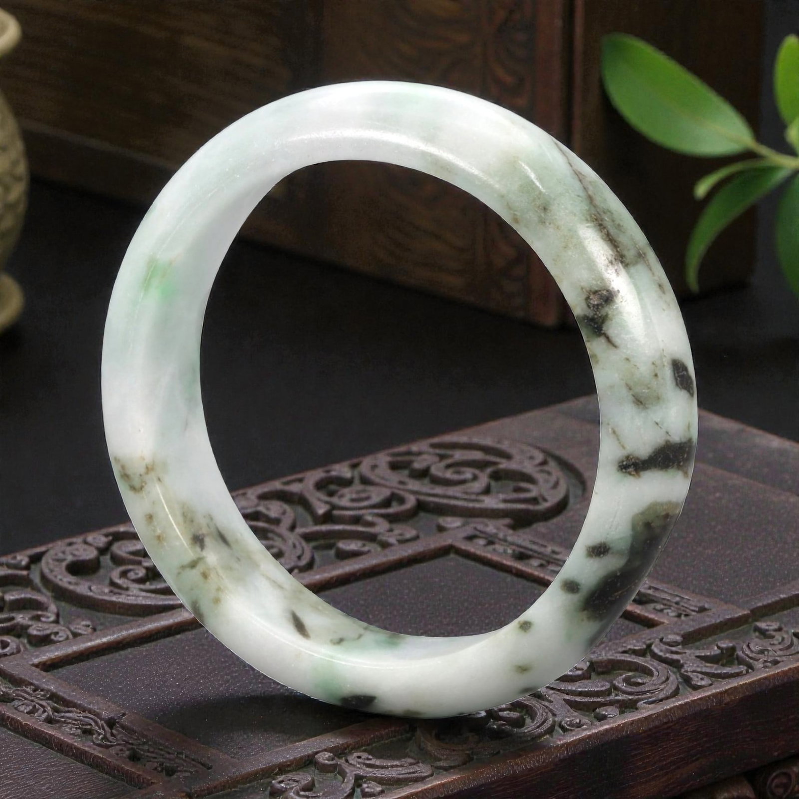 A Chinese Jadeite Jade Bangle Bracelet, 20th Century, 88ZK: Carved from jadeite jade with a pale white base and scattered dark green and black inclusions, the bangle is formed as a continuous circular band with a rounded cross-section. The surface is smoothly 