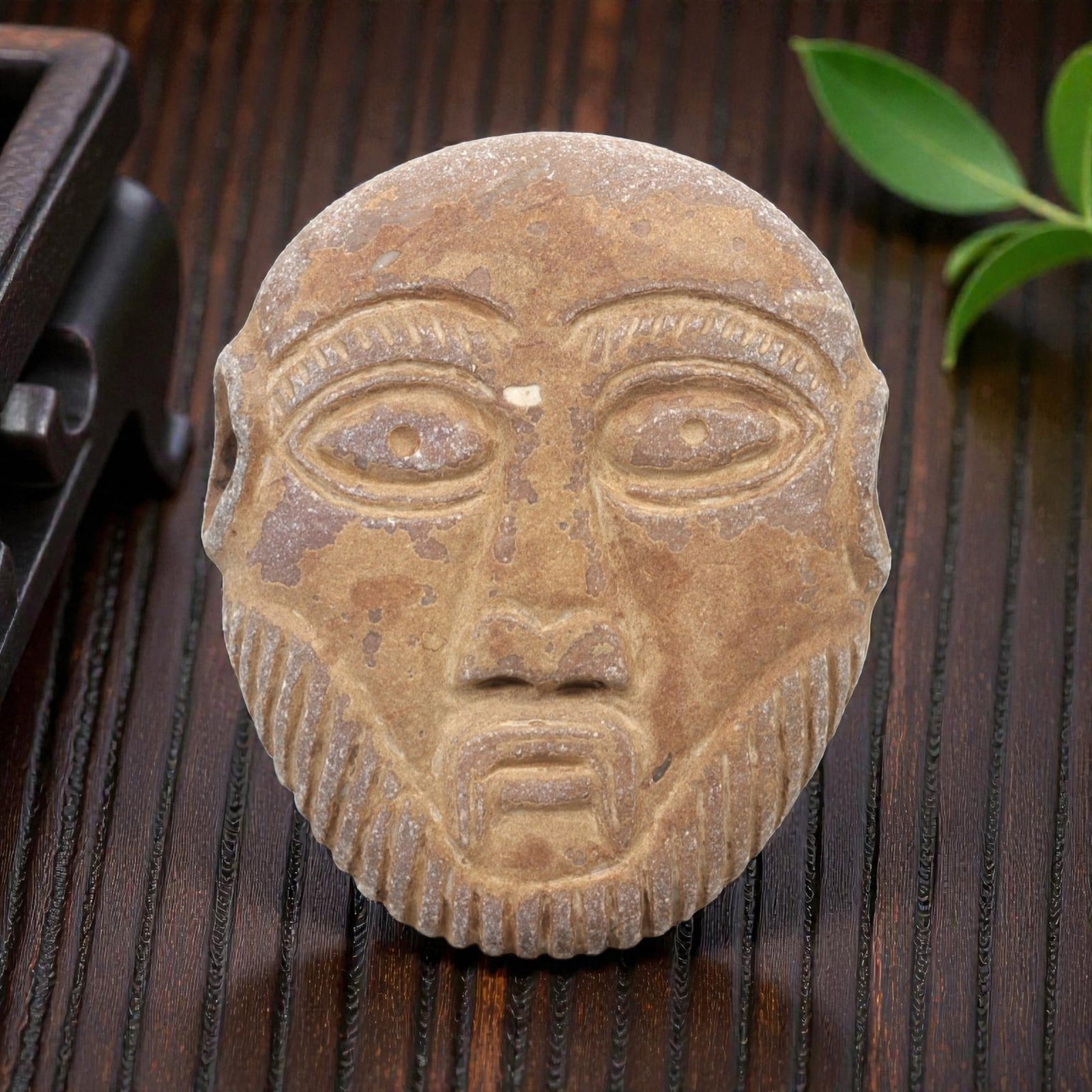 A Near Eastern Stone Maskette, 1st Millennium BCE, 29FL: Carved from brownish-tan stone with natural surface patination and minor chipping, the maskette is shaped as a rounded face with prominent, stylized features. The eyes are large and almond-shaped, eac