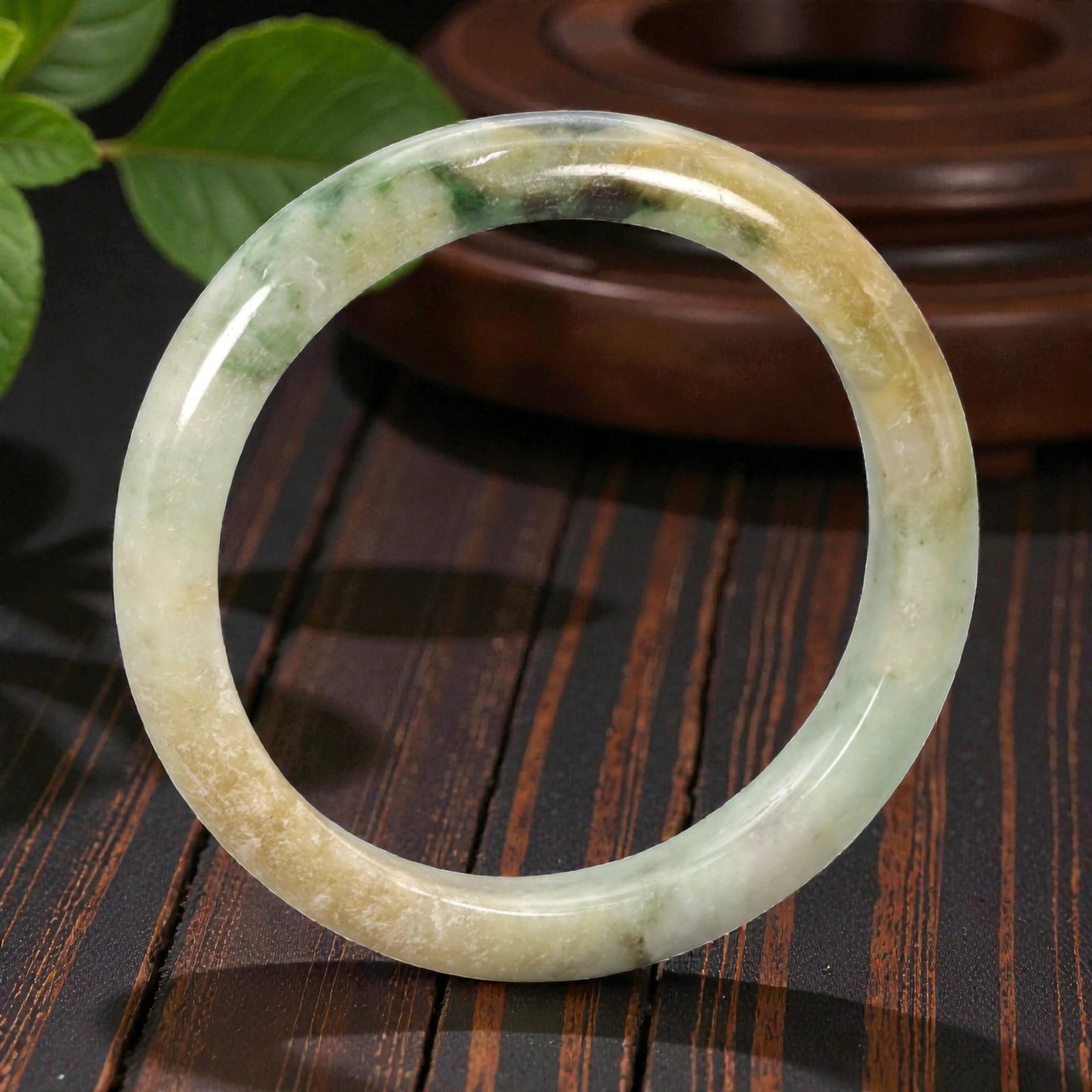 A Chinese Jadeite Jade Bangle Bracelet, 20th Century, 83ZK: Carved from jadeite jade with natural variegated coloration, including pale green, yellowish-brown, and darker green inclusions, the bangle is formed as a continuous circular band with a rounded, slig