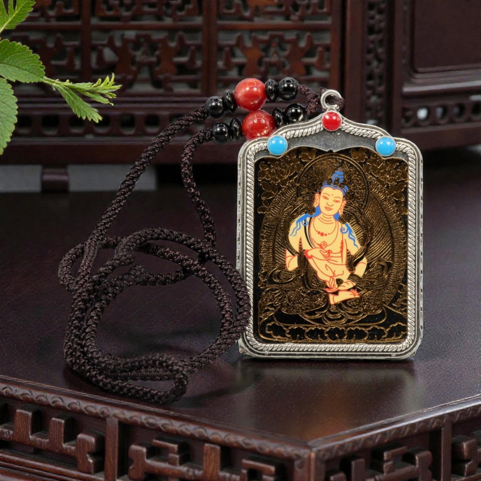 A Tibetan Metal and Painted Amulet, 20th Century, 48YC: The amulet is constructed with a rectangular metal frame housing a painted devotional image, suspended from a braided cord with applied beads. The central panel depicts a seated Buddhist figure shown 