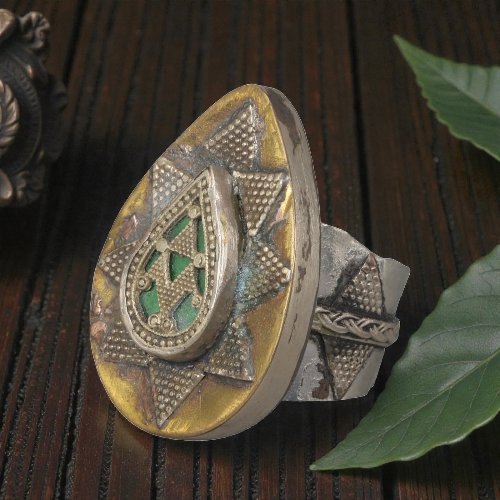 A Central Asian Silver Plated Teardrop Form Ring, Late 19th-Early 20th Century, 10AK: Crafted from silver plated metal with brass accents, featuring a wide, scalloped band. The teardrop-shaped bezel displays a central raised teardrop panel with green inlaid details and geometric openwo