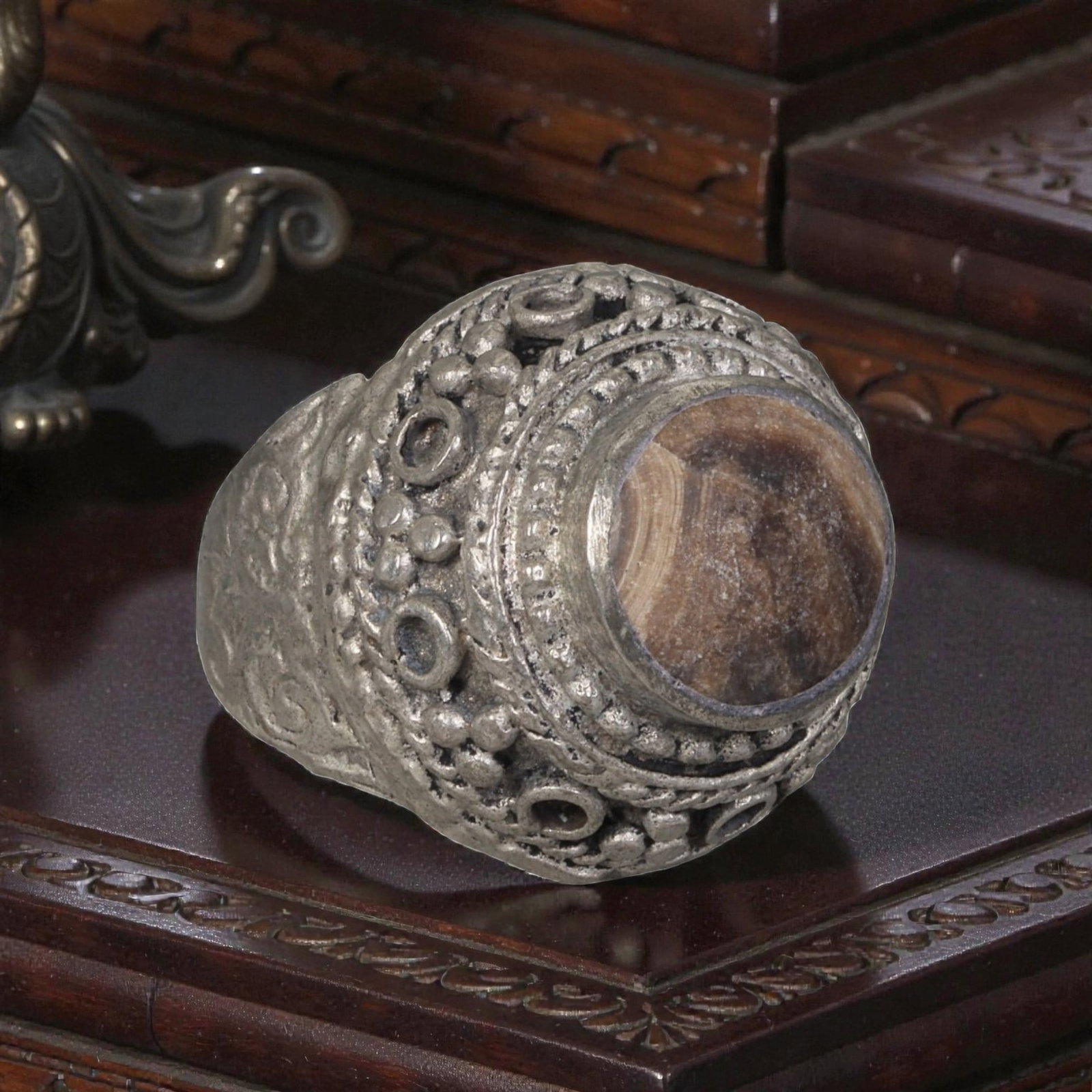 A Middle Eastern Silver Plated Banded Agate Ring, Late 19th-Early 20th Century, 30AK: Set with a round cabochon banded agate stone exhibiting natural layered brown and beige striations, the ring features an ornate domed bezel decorated with beaded granulation, twisted wirework, and emp