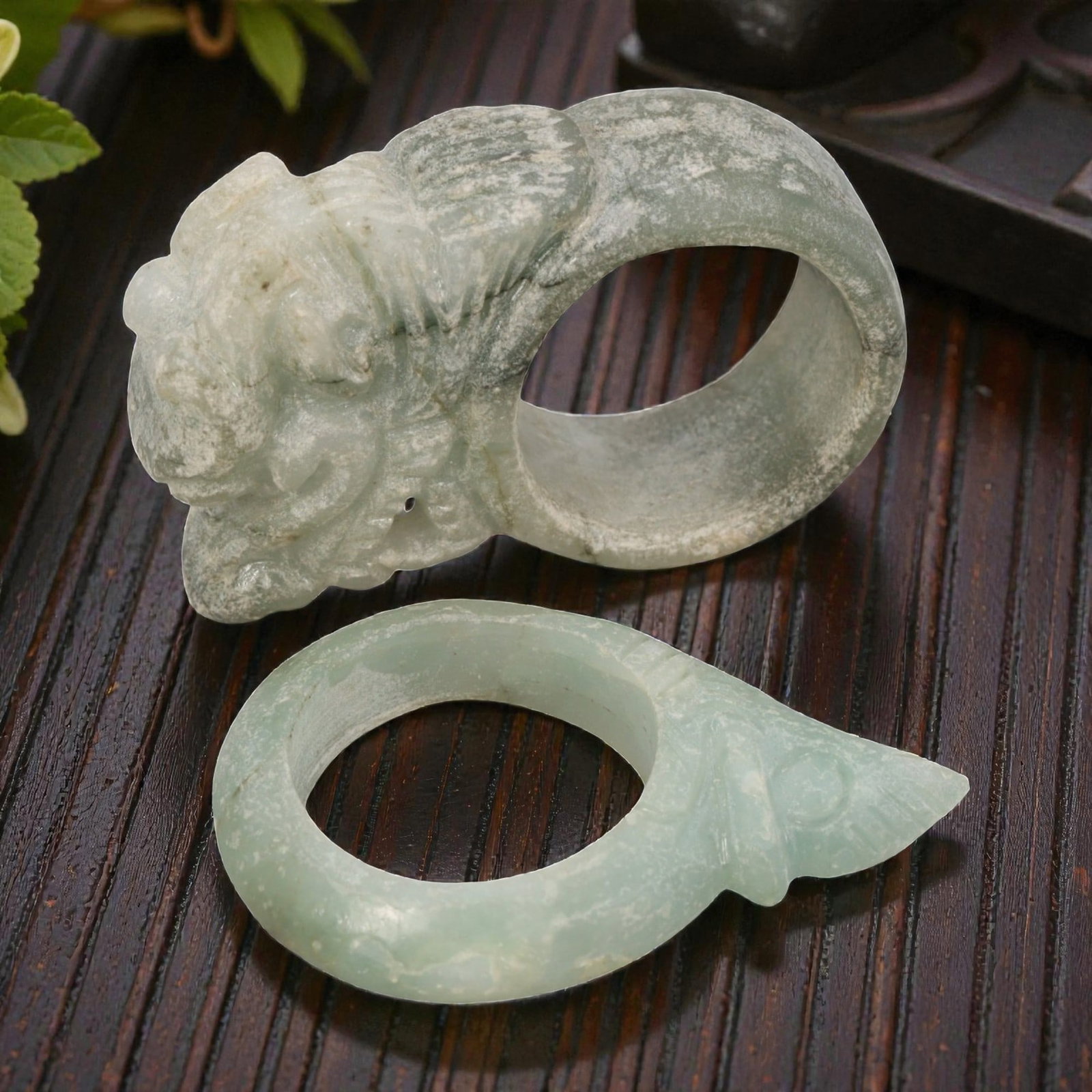 A Pair of Type A Jadeite Rings - Carved Animal, 16WS: This lot includes two type A jadeite rings, both carved from pale celadon-green jadeite with natural surface patina and mineral inclusions. The first ring features a detailed mythical guardian beast h