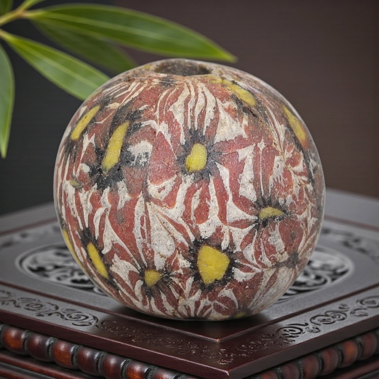 Antique Glass Bead - Islamic Jatim Red Floral Mosaic, 52YH: A spherical glass bead of Islamic Jatim type, crafted from polychrome mosaic glass. The surface features repeating stylized floral motifs with bright yellow centers and black outlines, set against a s