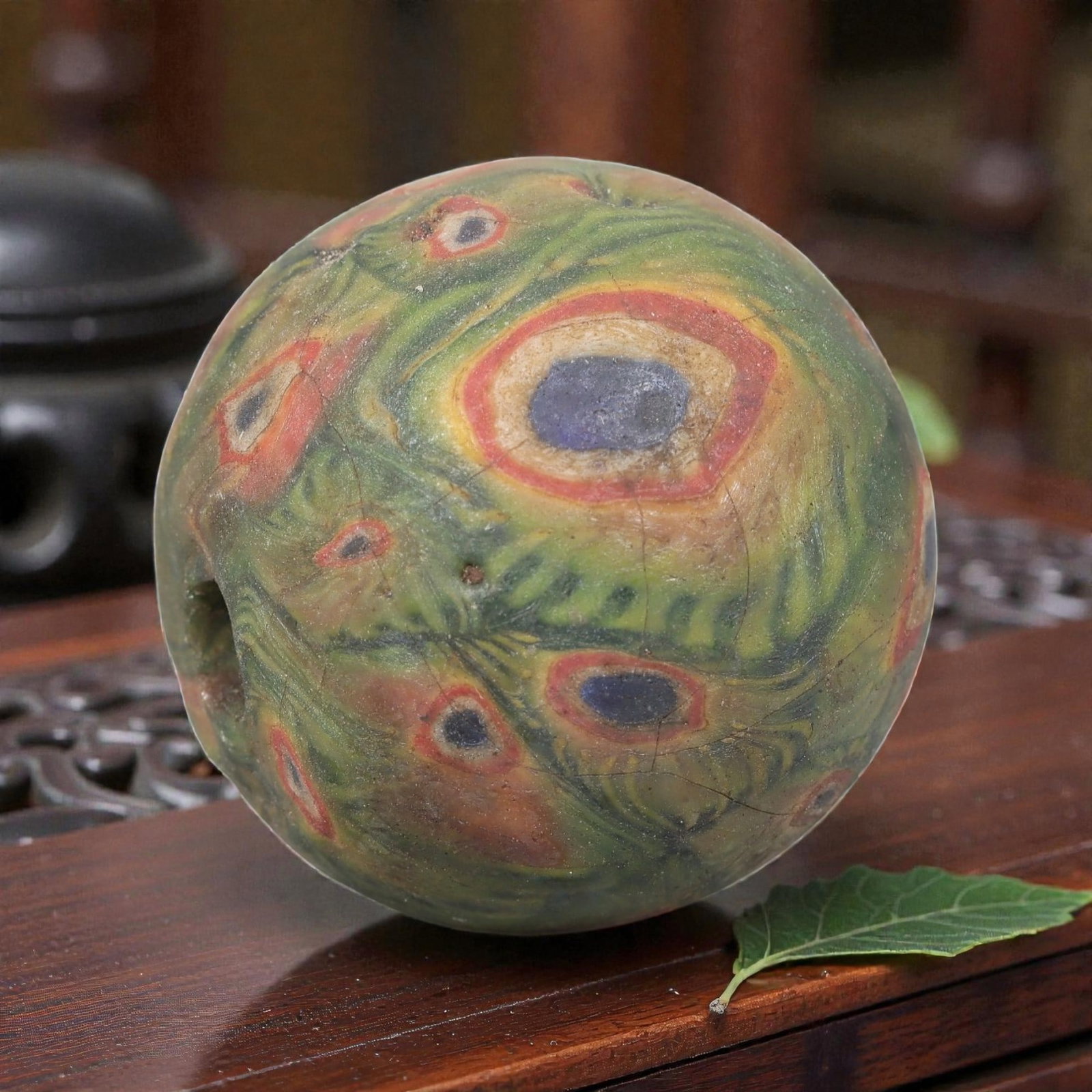 Antique Glass Bead - Islamic Jatim Green Peacock Eye Mosaic, 57AP: A spherical glass bead of Islamic Jatim type, crafted from polychrome mosaic glass. The surface features a striking peacock eye motif, with deep blue centers encircled by red and yellow rings, set aga