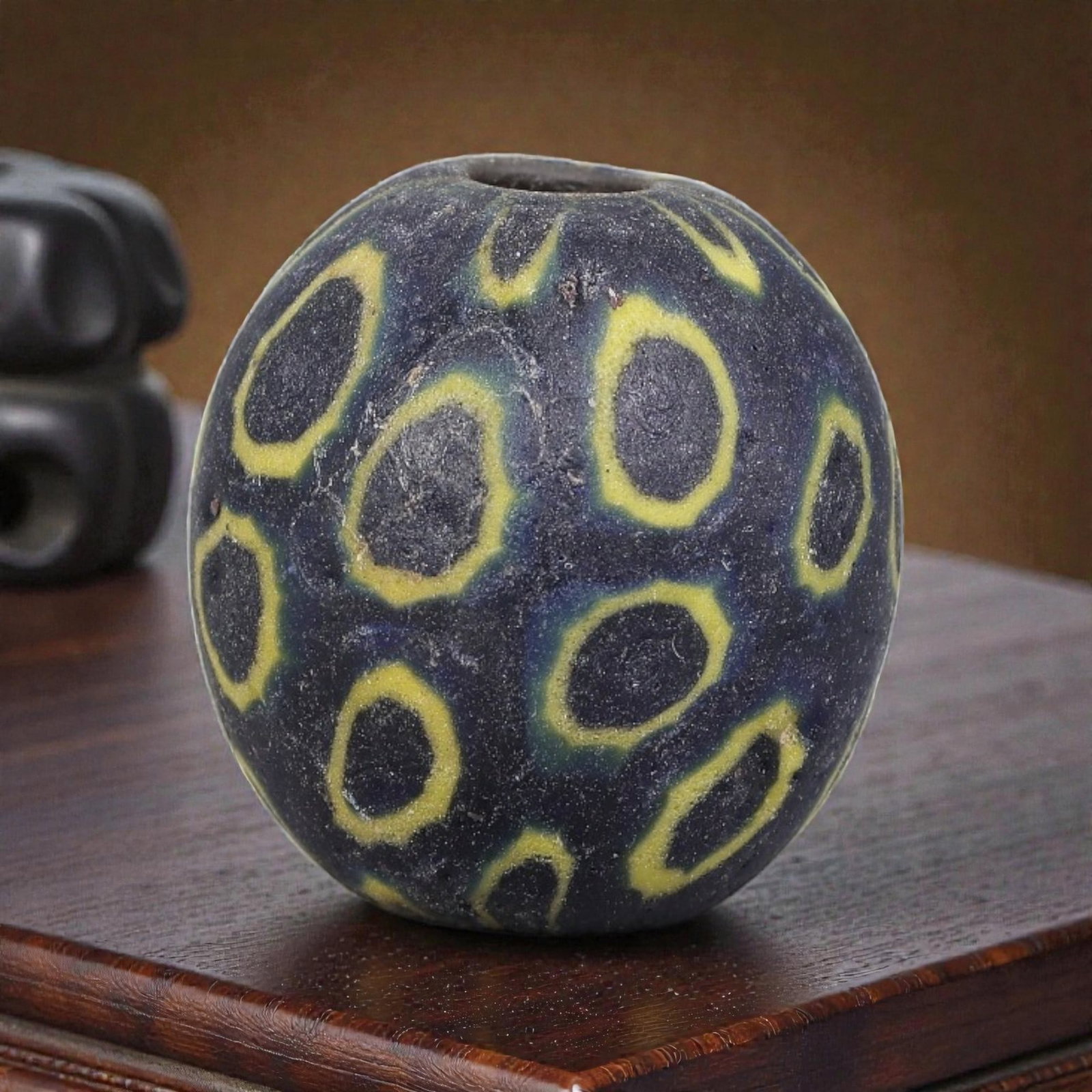 Antique Glass Bead - Islamic Jatim Indigo Yellow-Ring Eye Mosaic, 40YH: A spherical glass bead of Islamic Jatim type, crafted from polychrome mosaic glass. The surface features irregular eye motifs with dark indigo centers framed by bright yellow borders, set against a te