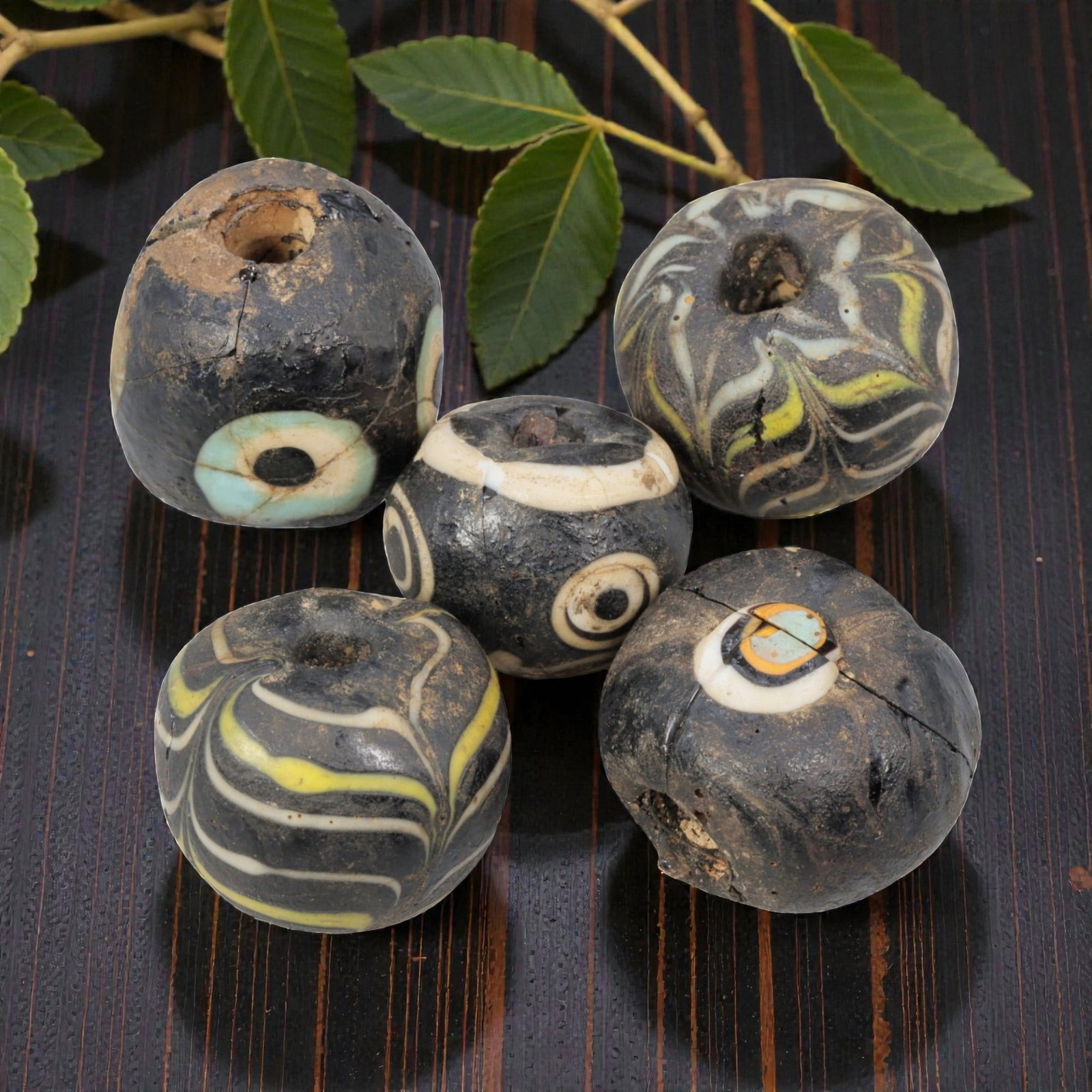 Lot of 5 Antique Islamic Glass Beads, Circa 9th-14th Century, 73AD: Comprising five beads ranging from 1.5 cm (5/8 in.) to 2 cm (3/4 in.), each formed from dark base glass with varied decorative motifs. Two beads feature feather-patterned trails in white, yellow, and 