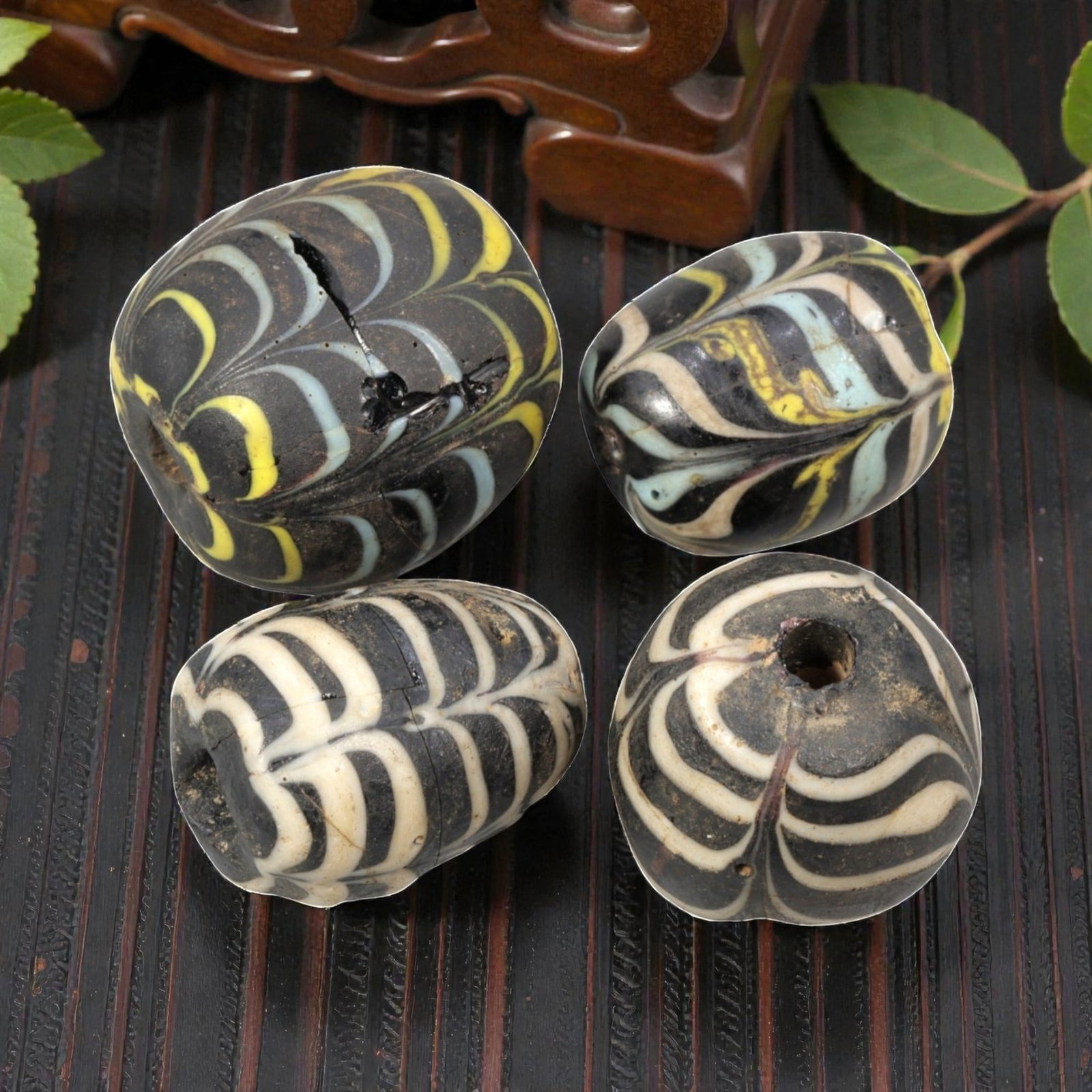 Lot of 4 Natural Islamic Feather-Patterned Glass Beads, Circa 9th-14th Century, 68AD: A group of four glass beads, each with a black base decorated with feather-patterned trailing in white, light blue, and yellow glass. The beads are barrel-shaped with central perforations, showing sig