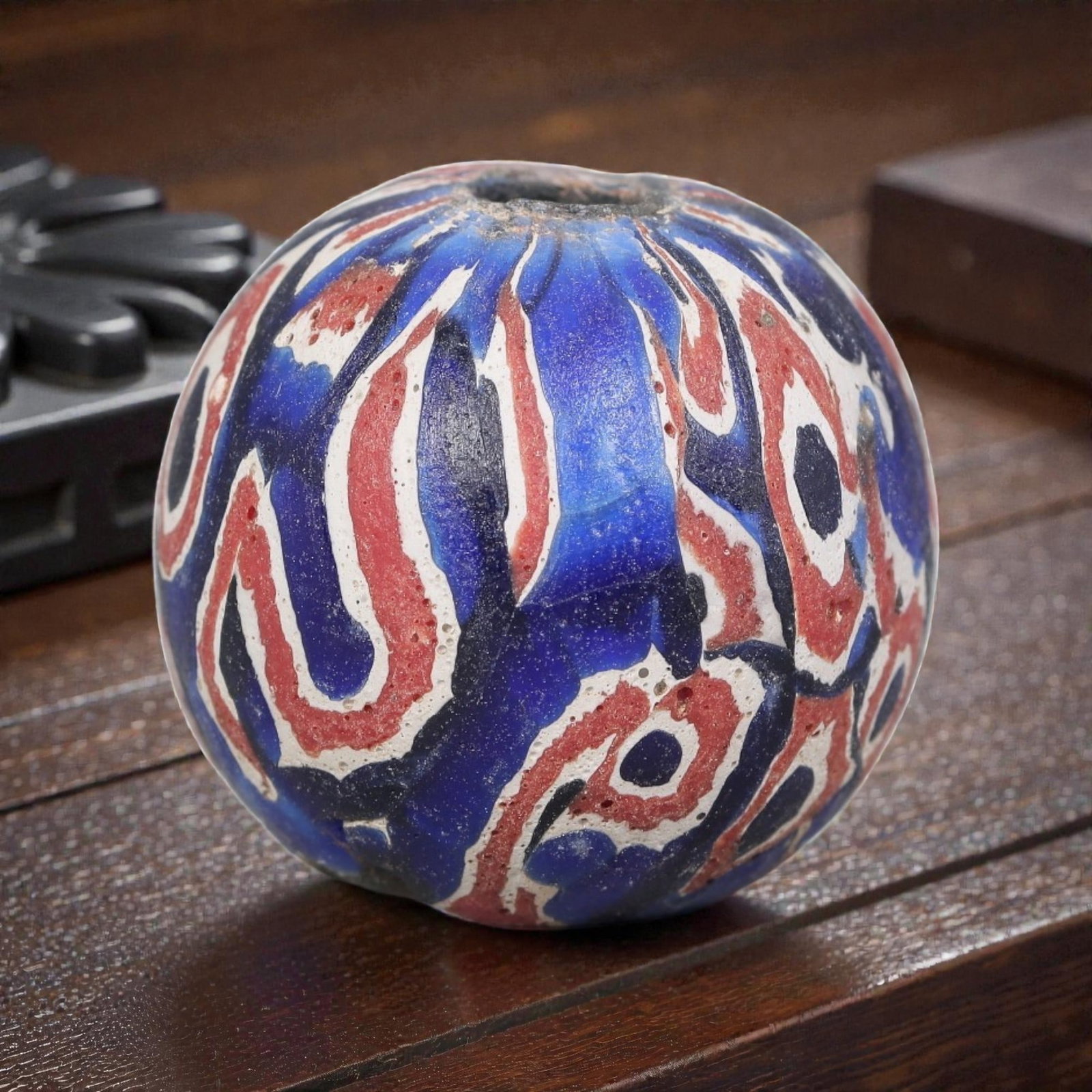 Antique Glass Bead - Islamic Jatim Red & Blue Pattern, 74AP: A spherical glass bead of Islamic Jatim type, crafted from polychrome mosaic glass. The surface features an intricate red, white, and blue pattern, with bold, curvilinear motifs set against a deep cob