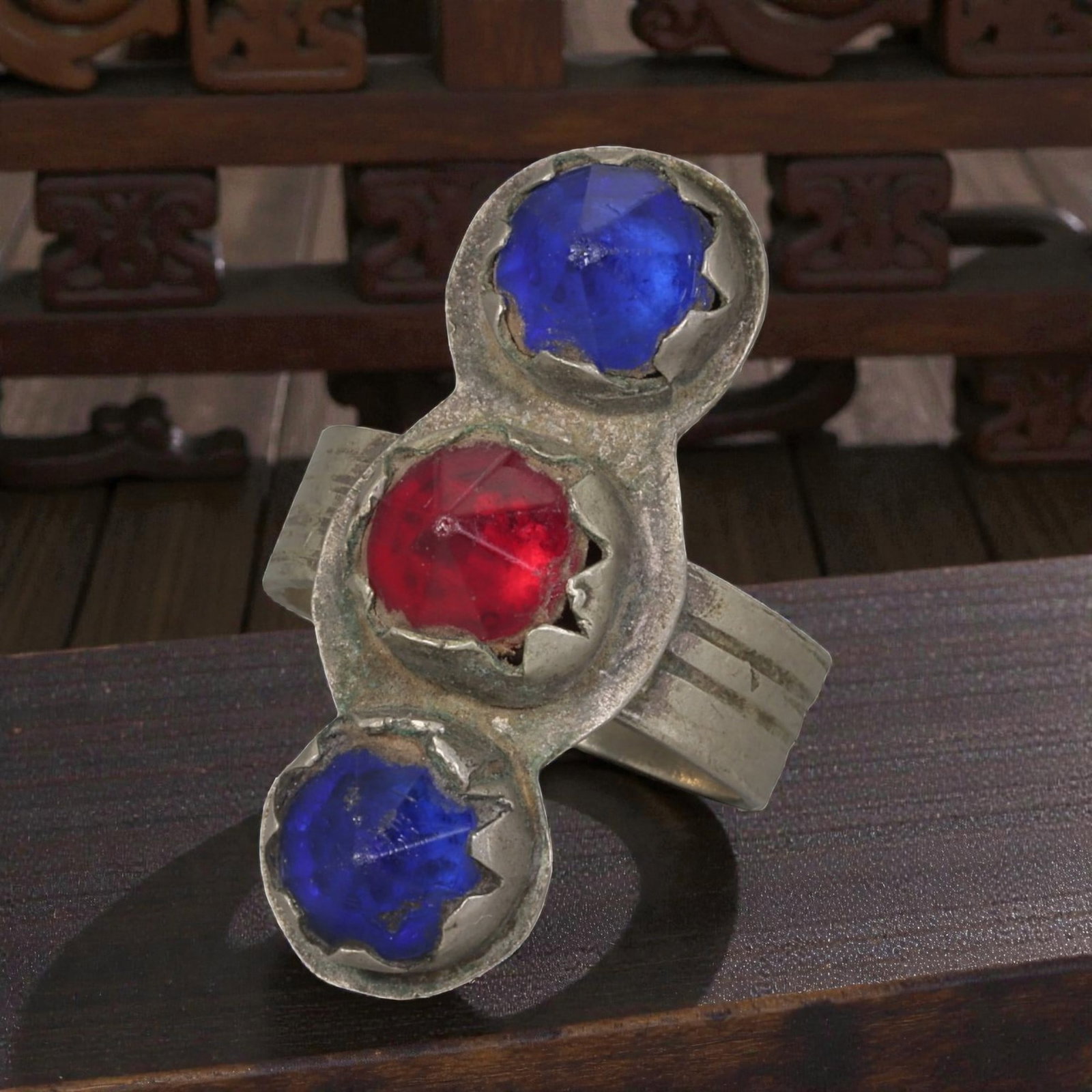 A Berber Silver Three-Stone Ring, Late 19th-Early 20th Century, 38AK: Crafted from silver, featuring a wide band with parallel incised linear detailing. The vertical bezel holds three faceted stones: two blue and one red, set in scalloped bezels. The metal surface shows