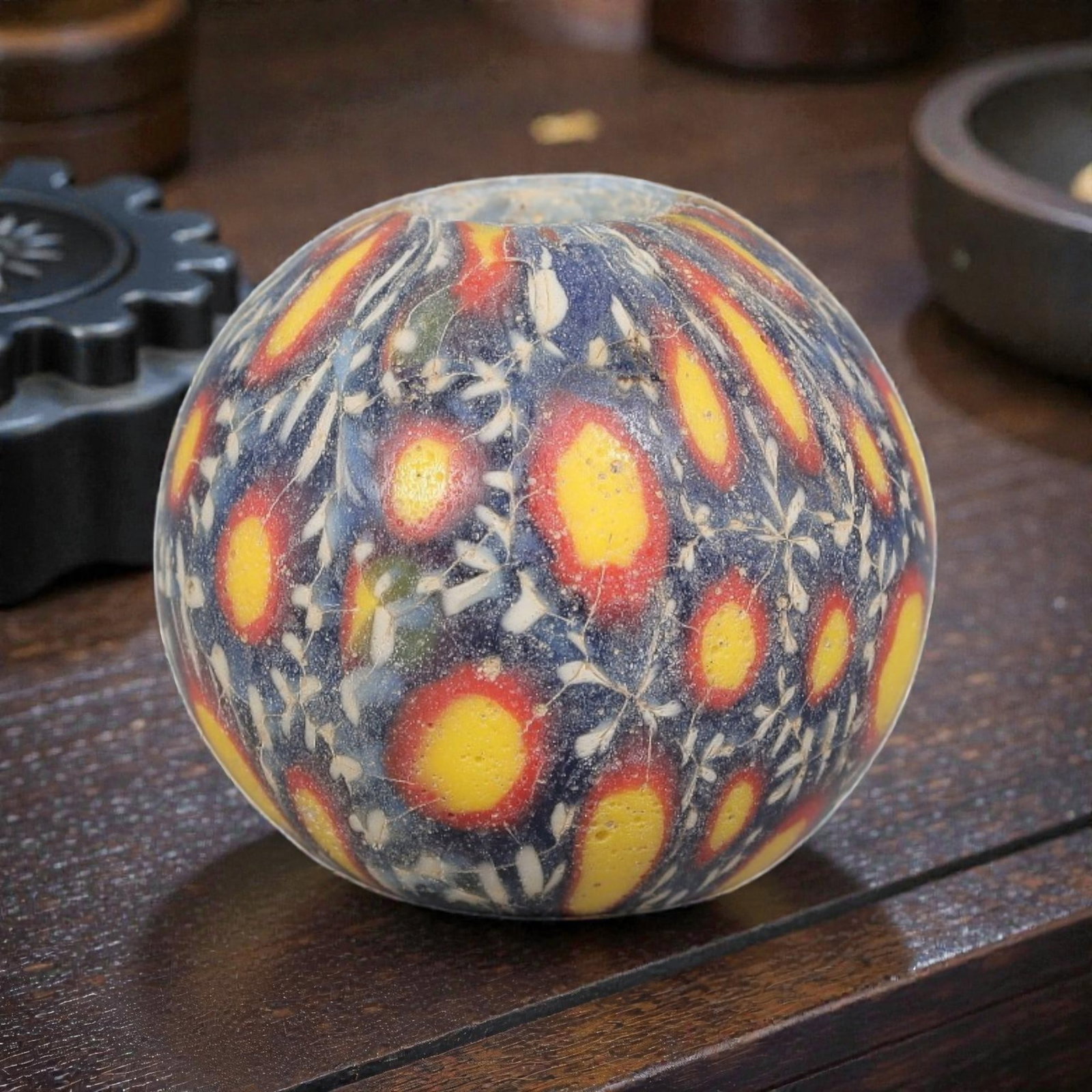 Antique Glass Bead - Islamic Jatim Multicolor Peacock Eye Mosaic, 57YH: A spherical glass bead of Islamic Jatim type, crafted from polychrome mosaic glass. The surface features repeating peacock eye motifs with bright yellow centers and red borders, set against a deep blu