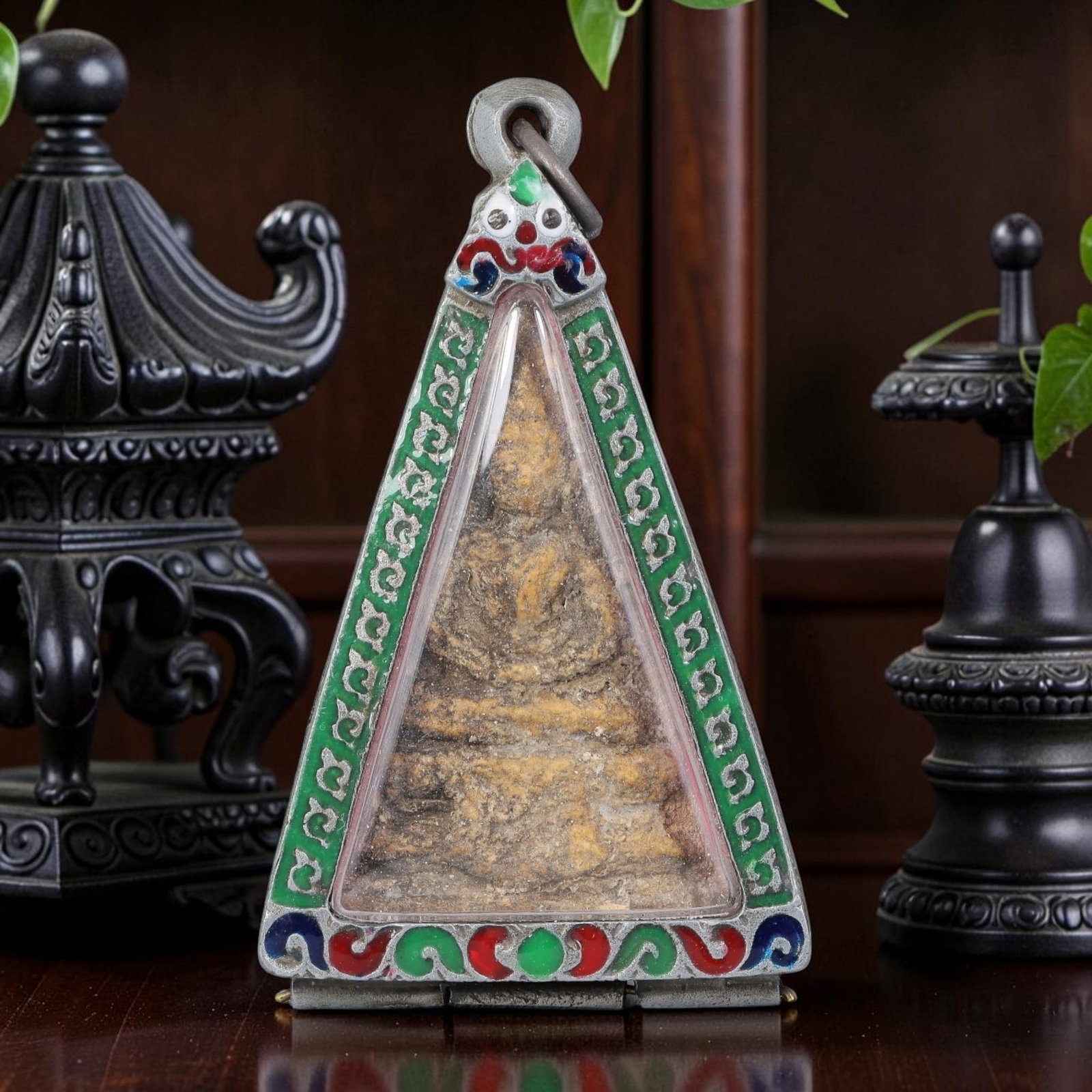 Thai Buddha Amulet - Triangular Enamel Silver Tone Pendant, 49AP: A Thai Buddhist amulet featuring a seated Buddha figure, housed in a triangular silver-tone metal casing with colorful enamel detailing. The glass-fronted enclosure protects the aged Buddha plaque, wh