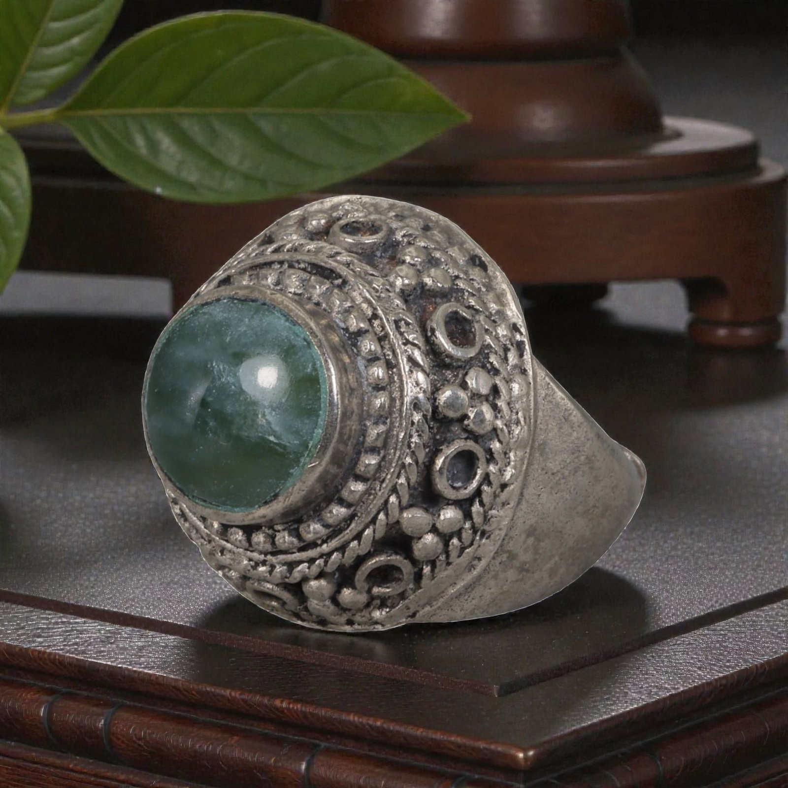 A Middle Eastern Silver Plated Green Agate Ring, Late 19th-Early 20th Century, 17AK: Set with a round cabochon green agate stone exhibiting natural mottled green tones, the ring features an ornate domed bezel decorated with beaded granulation, twisted wirework, and empty circular sett