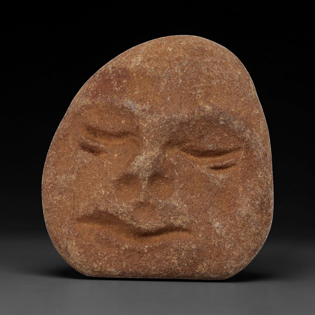Antique Medieval Carved Stone Maskette, 82AN: Carved from a single piece of weathered brown stone, this maskette depicts a stylized human face in relief. The figure features simplified, closed eyes, a prominent nose, and subtle lips, with the sto