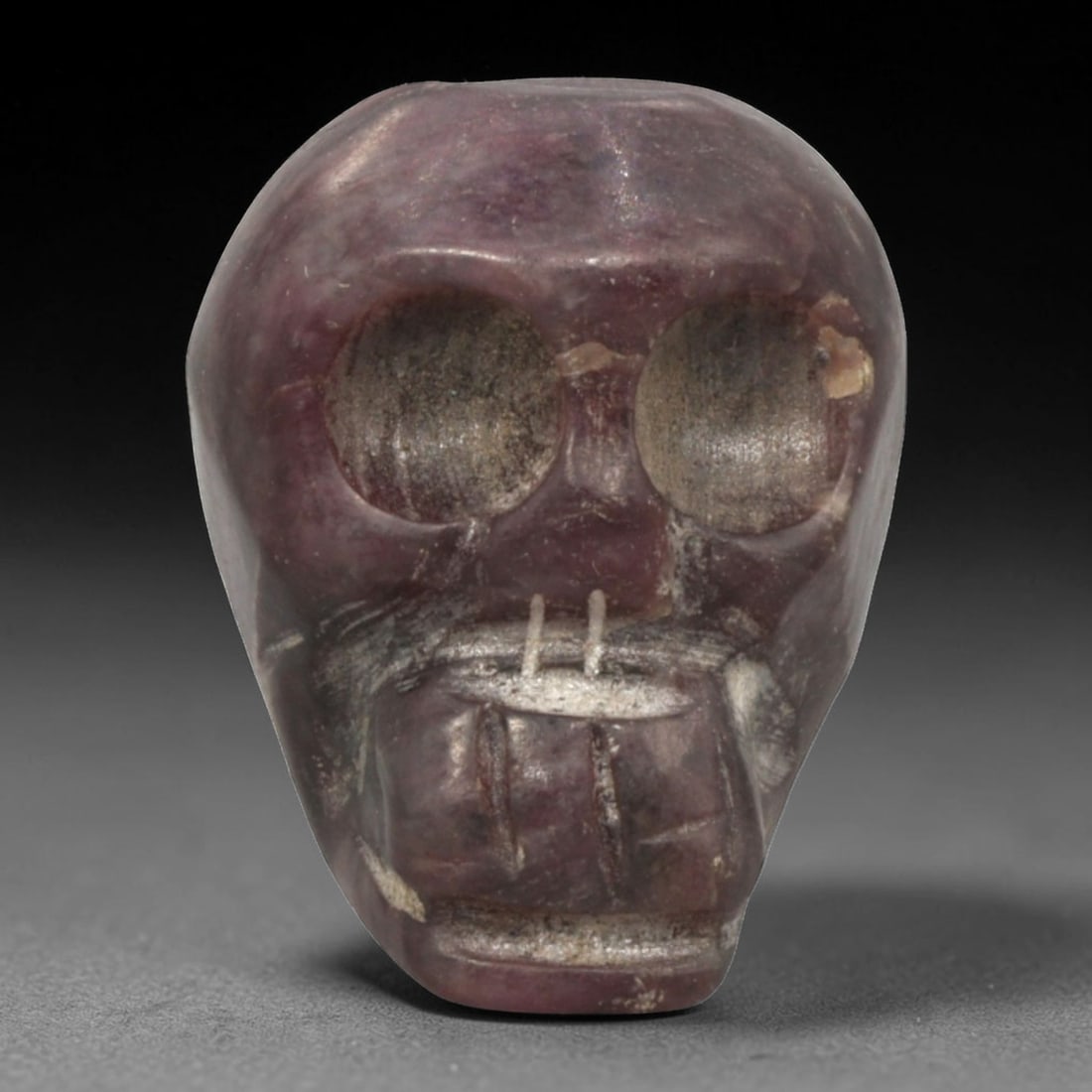 Antique Tibetan Purple Stone Skull Bead, 1AD: Carved from natural purple stone with a rich variegated hue and crystalline texture, the bead is shaped as a stylized human skull. The front face features large, hollowed eye sockets, a simplified nas