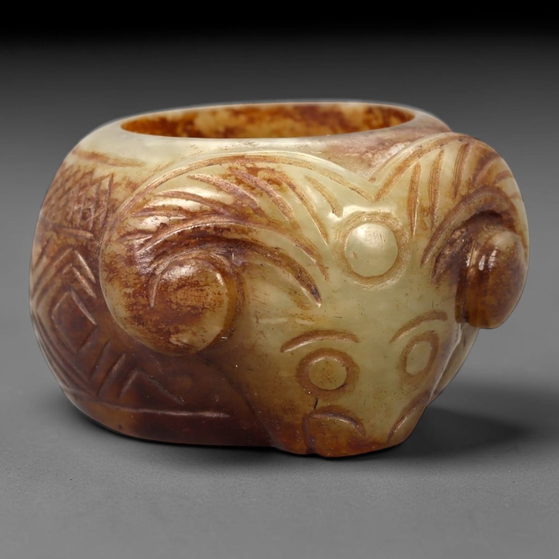 A Chinese Nephrite Jade Ram-Head Ring, 14FA: Carved from mottled celadon and russet-toned nephrite jade with natural color variation and areas of translucency, the ring is formed with a sculptural ram's head projecting from the bezel. The head i