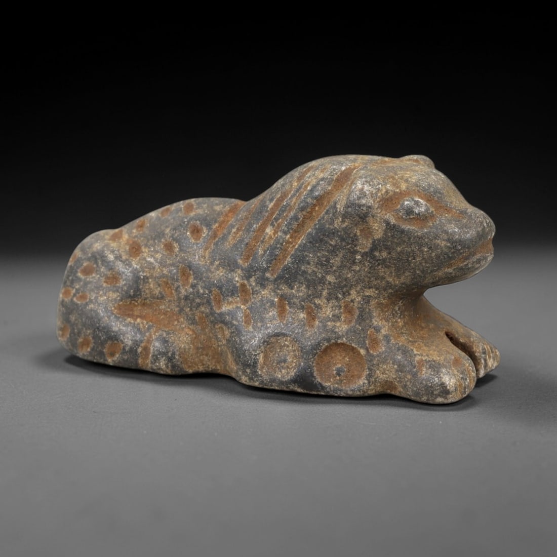 Ancient Stone Zoomorphic Amulet - Near Eastern Feline Figure, 12AM: This amulet is carved from dark stone, depicting a recumbent feline in a crouched posture, with incised linear and circular motifs defining the mane, spots, and facial features. The piece reflects the