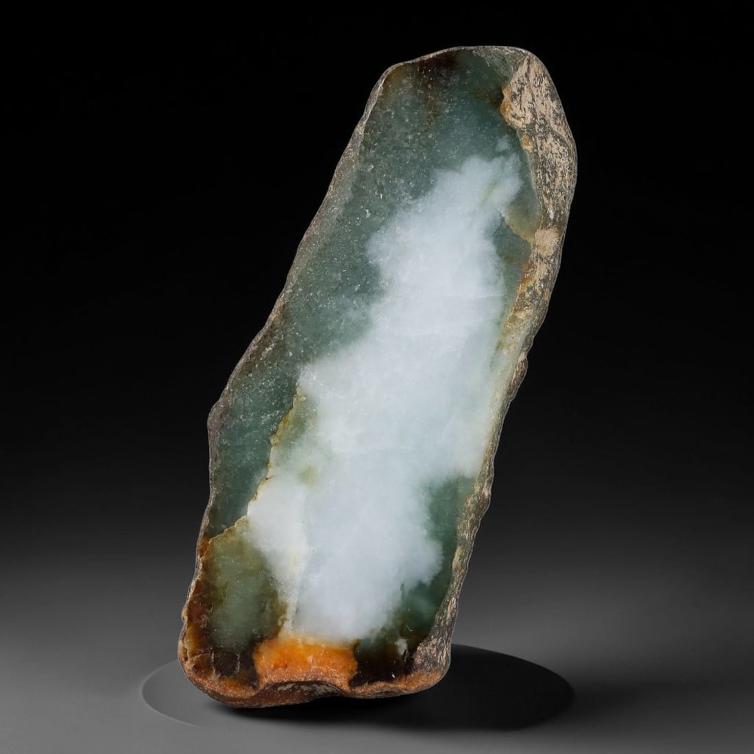 225ct Natural Type A Burmese Central White Cloud Pattern Teal Jadeite Jade Sliced Rough, 5FA: This specimen is an elongated sliced rough slab of untreated Type A Burmese jadeite, retaining naturally weathered multi-toned brown and earthy rind along its irregular perimeter edges, with a strikin