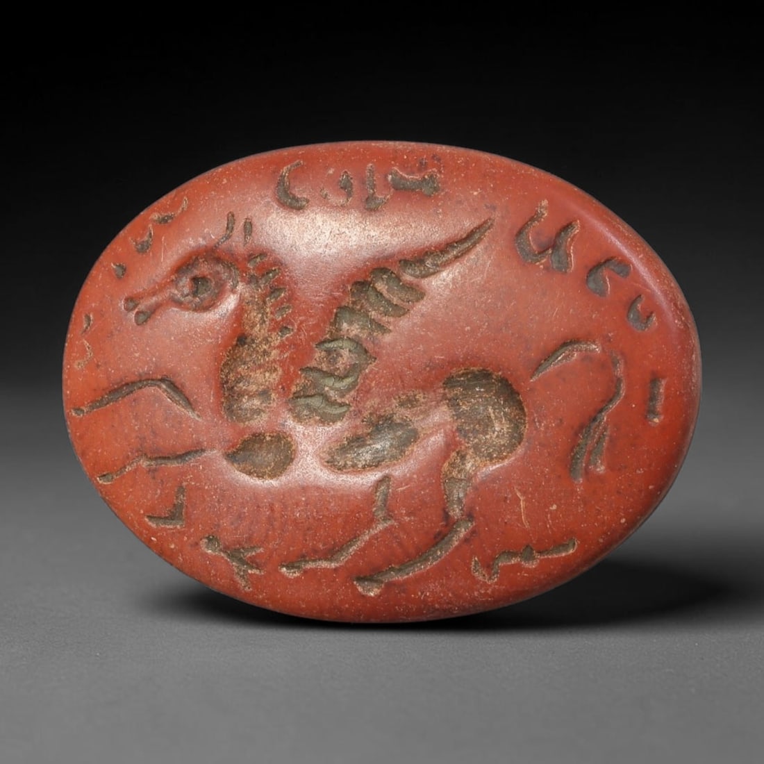 Ancient Sasanian Persian Red Jasper Winged Horse Intaglio Seal Stone (224-651 CE), 85AE: Hand-carved oval red jasper intaglio seal from Sasanian Persian civilisation, engraved with a dynamic winged horse motif encircled by stylised Pahlavi script inscriptions. The recessed carved lines ho