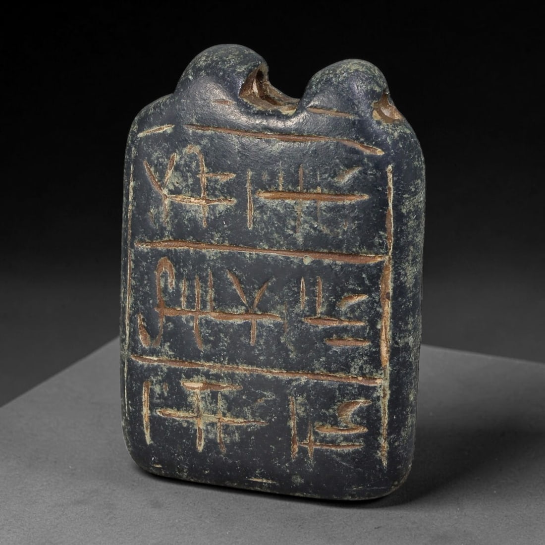 Ancient Egyptian Dark Faience Hieroglyphic Tablet Amulet (664-30 BCE), 84AE: Rectangular dark glazed faience pendant tablet with twin bulbous suspension loops drilled for cord threading. The front surface bears three horizontal registers of incised Egyptian hieroglyphic script