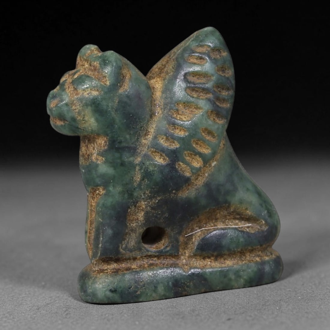 Ancient Western Asiatic Recumbent Beast Zoomorphic Amulet (1000-600 BCE), 26AB: Hand-carved mottled dark green serpentine pendant shaped as a seated feline beast set upon an integrated flat rectangular base. Detailed incised oval indentations adorn its raised wing-like crest, whi