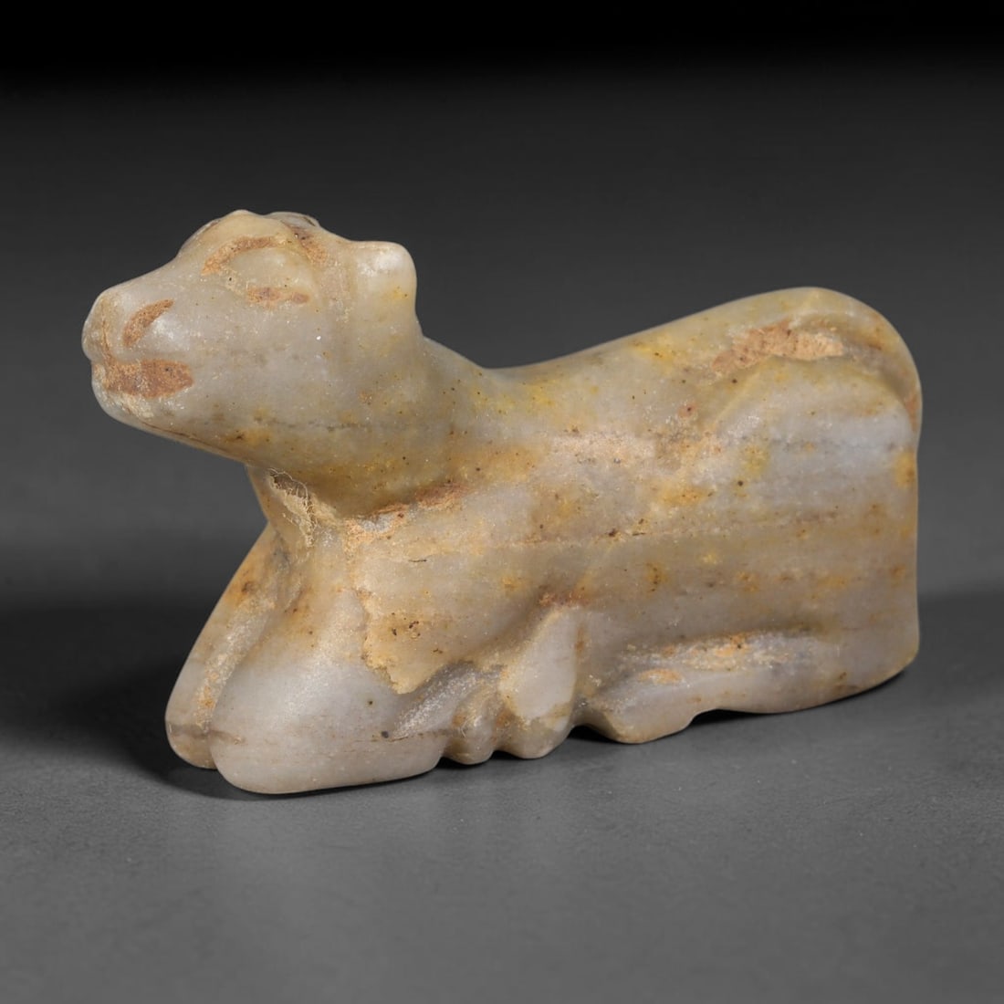 Ancient Near Eastern Pale Banded Calcite Recumbent Cow Amulet (800-300 BCE), 42AB: Hand-carved miniature talisman from creamy pale banded calcite, shaped as a reclining cow with stylised simplified facial incisions for eyes, nose and mouth. Warm ochre burial staining spreads across 