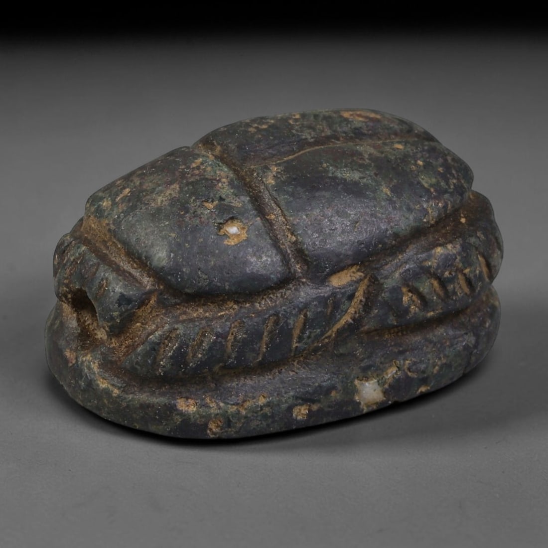 Ancient Egyptian Late Period Dark Hardstone Scarab Amulet (664-30 BCE), 64AB: Hand-carved dark mottled hardstone scarab funerary talisman, featuring incised linear grooves detailing wing casings and segmented legs. Thick earthy burial patina coats recessed carving lines, scatte