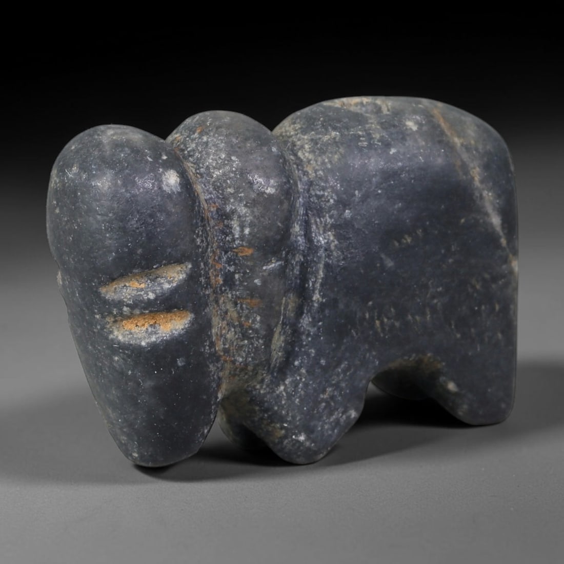 Ancient Western Asiatic Dark Grey Stone Stylized Elephant Zoomorphic Amulet (1000-600 BCE), 18AB: Hand-carved matte dark grey hardstone pendant shaped as a simplified standing elephant with large rounded ears, elongated trunk and minimal linear incisions for facial detailing. Faint rust-hued buria