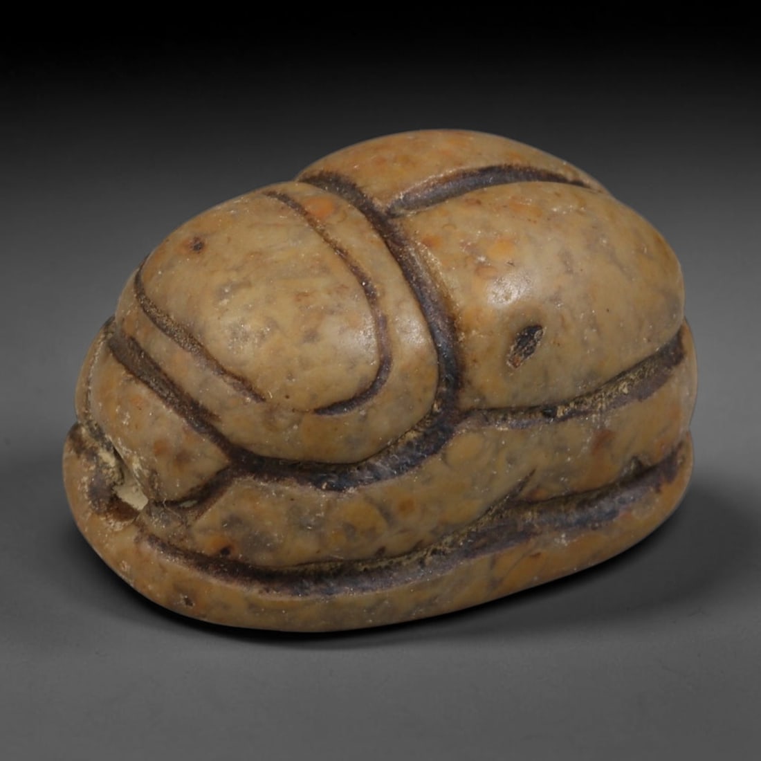 Ancient Egyptian Late Period Mottled Tan Hardstone Scarab Amulet (664-30 BCE), 48AB: Hand-carved domed scarab talisman fashioned from warm mottled beige hardstone, defined by deep incised grooves separating the head, thorax and paired wing casings. Dark aged soil patina fills every ca