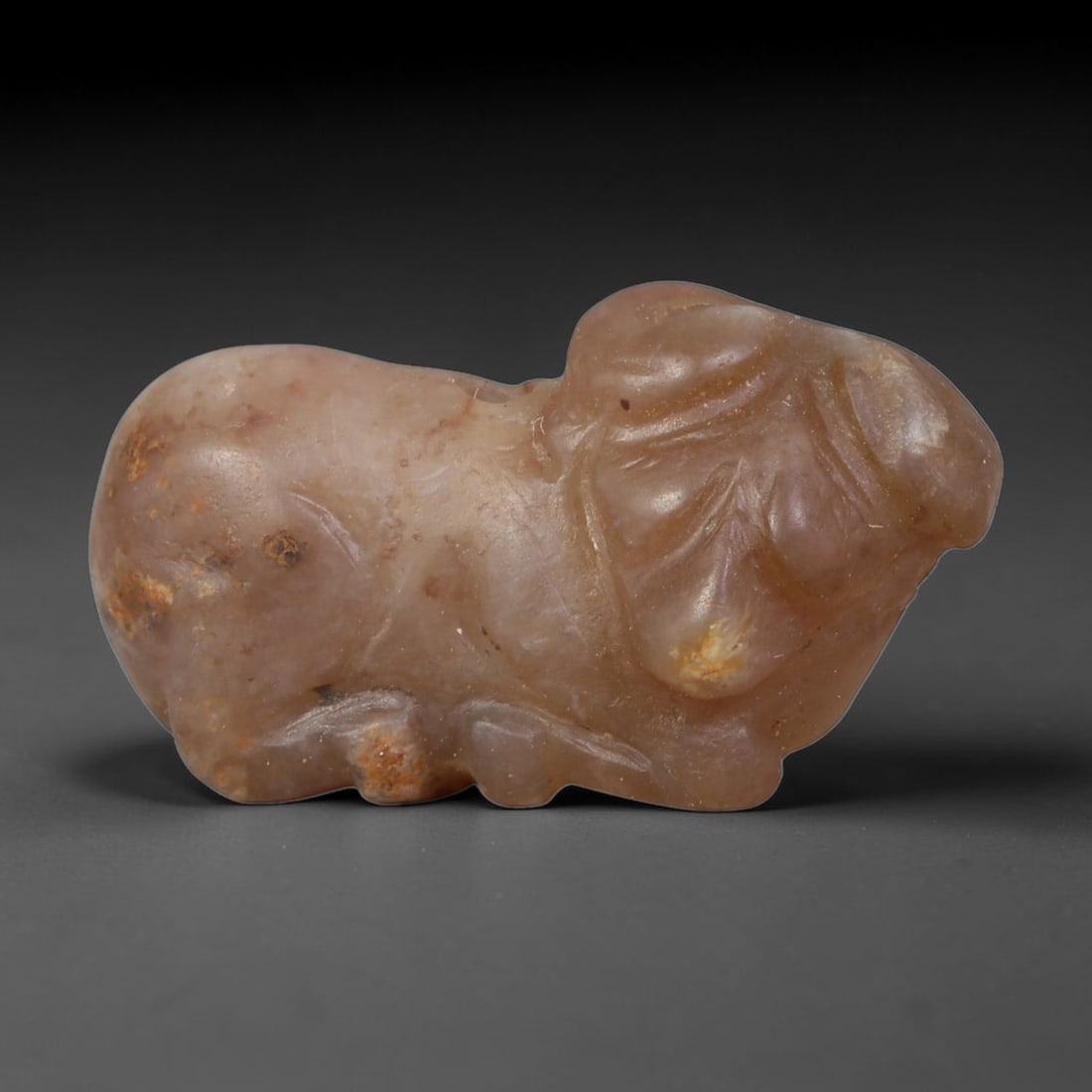 Ancient Western Asiatic Pinkish Agate Recumbent Ram Zoomorphic Amulet (1000-600 BCE), 29AB: Hand-carved semi-translucent pale blush agate pendant shaped as a resting ram with simplified incised detailing for curled horns, facial folds and folded legs. Warm rust-coloured mineral patina flecks