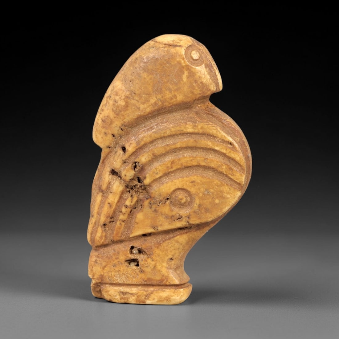 Ancient Bone Bird Amulet - Near Eastern Figurine, 84AL: Carved from tan bone, this amulet depicts a stylized avian form with a rounded head, incised eye, and curved beak. The body features concentric incised linear patterns creating a layered, feather-like