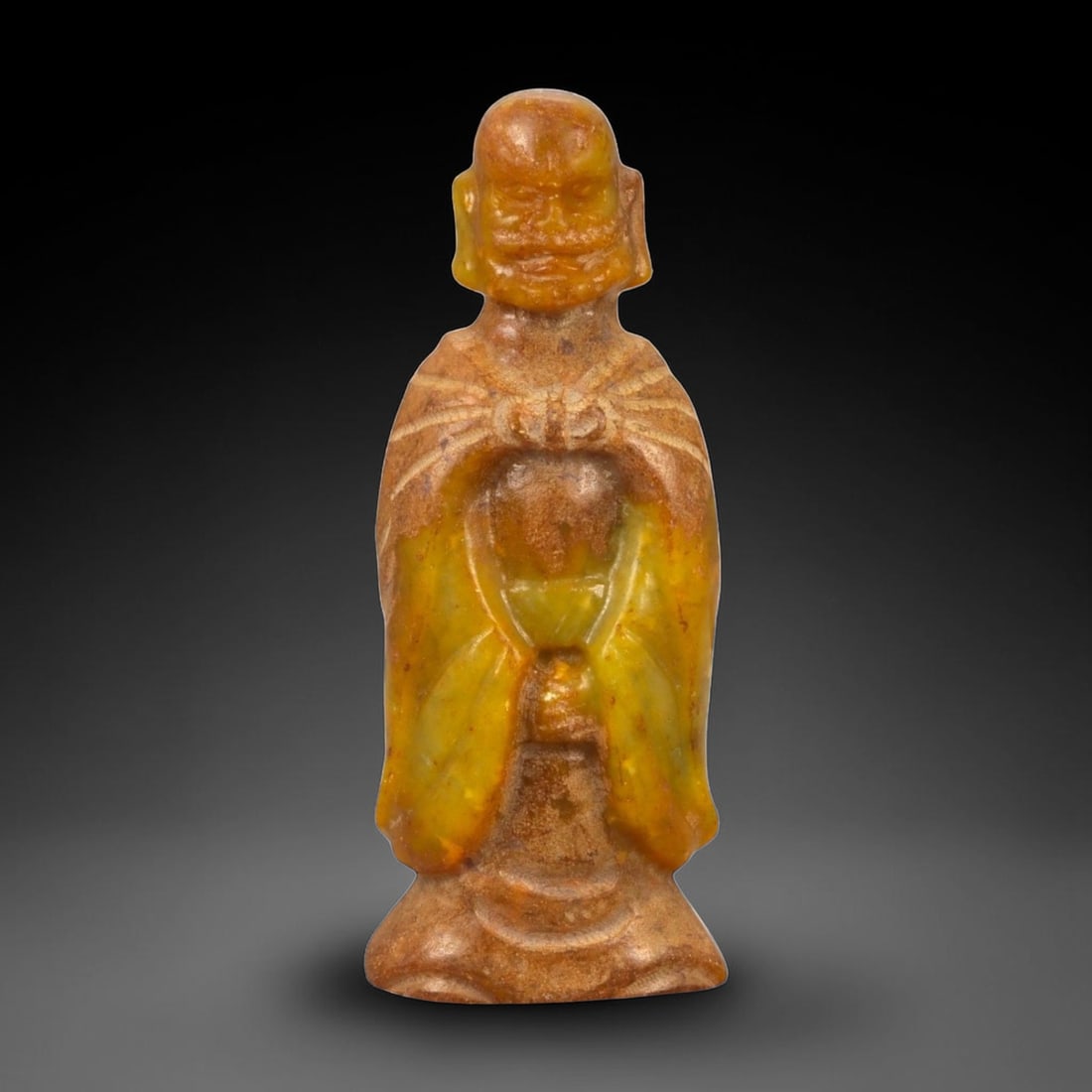A Chinese Jade Figure of a Standing Monk, 20th Century, 65FB: Carved from translucent yellow to brown-toned jade with areas of darker inclusions and natural surface variation, the figure represents a standing male monk shown frontally. The head is bald, with sim