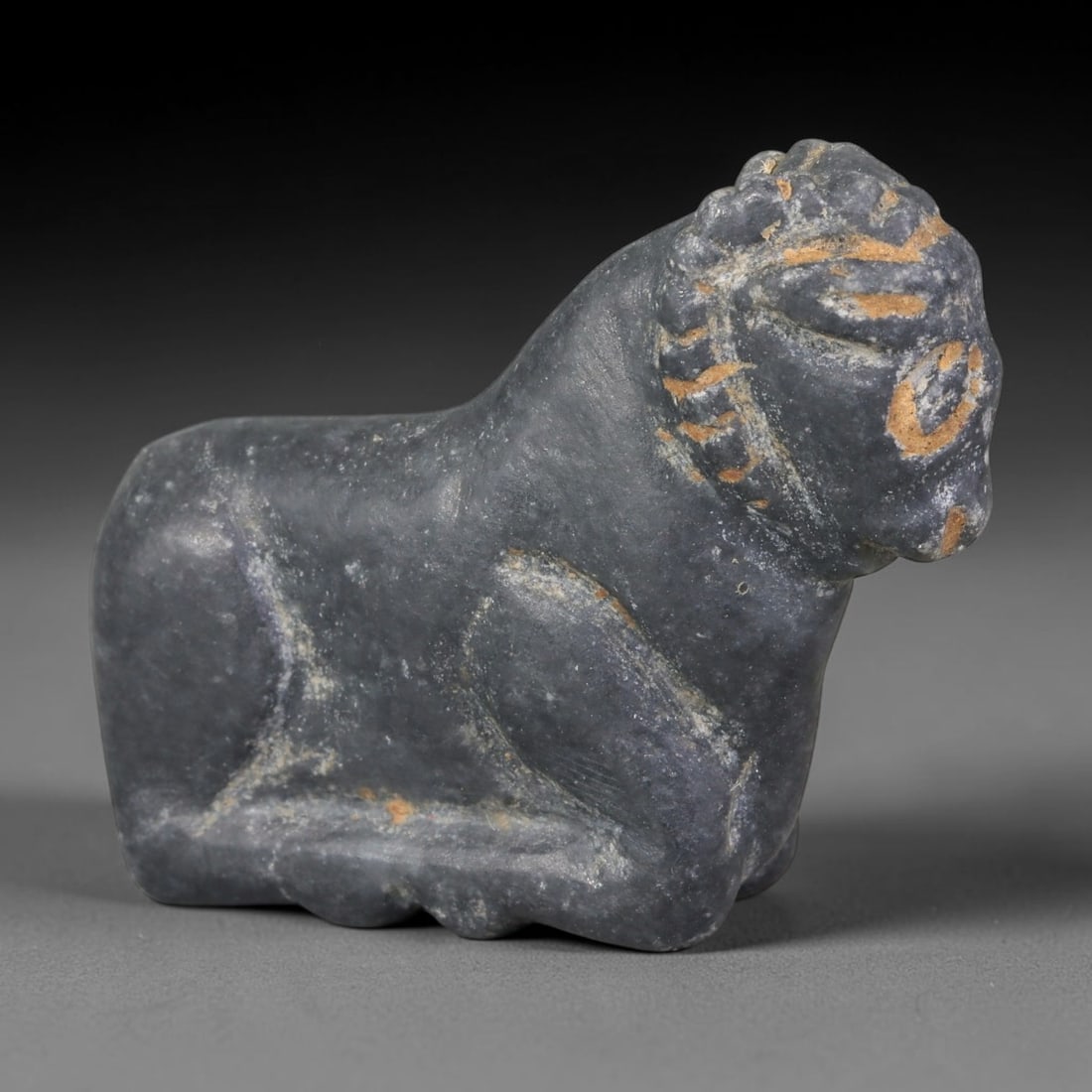 Ancient Near Eastern Dark Grey Steatite Recumbent Ram Amulet (800-300 BCE), 43AB: Hand-carved miniature talisman crafted from smooth dark grey steatite, modelled as a reclining ram with stylised curled spiral horns and simple incised facial details. Warm ochre burial patina settles