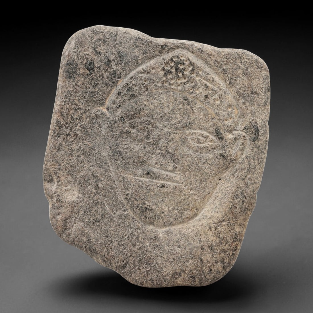 Ancient Sino-Siberian Carved Stone Plaque, 52AG: A carved stone plaque of irregular form, featuring an incised human face with stylized features, including defined eyes, nose, mouth, ears, and a decorated headdress. The stone surface is weathered wi