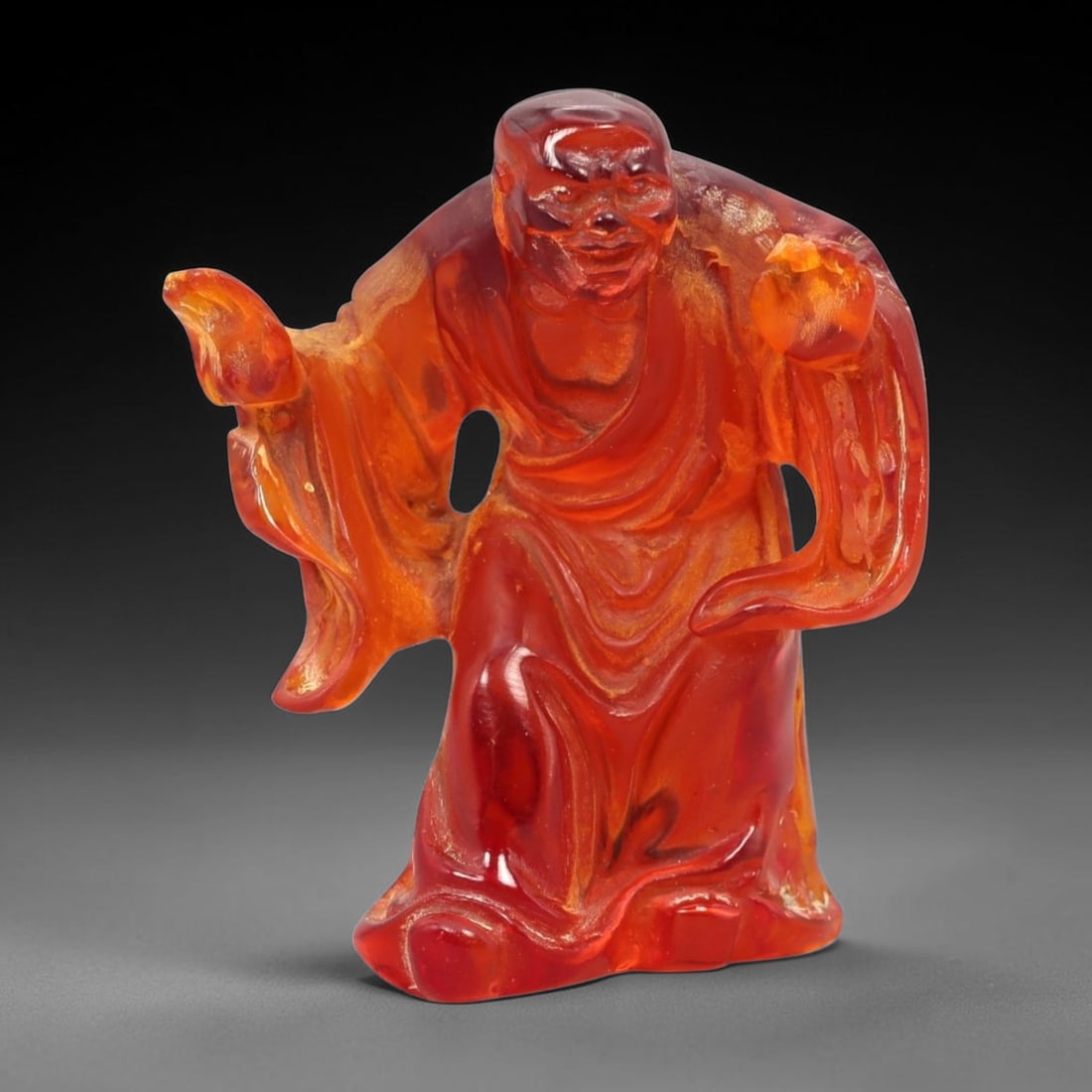 A Chinese Amber-Resin Figure of a Luohan, 42FH: Cast or carved from translucent amber-colored red resin with internal tonal variation, scattered inclusions, and minor entrapped bubbles, the figure represents a standing luohan shown in an animated p