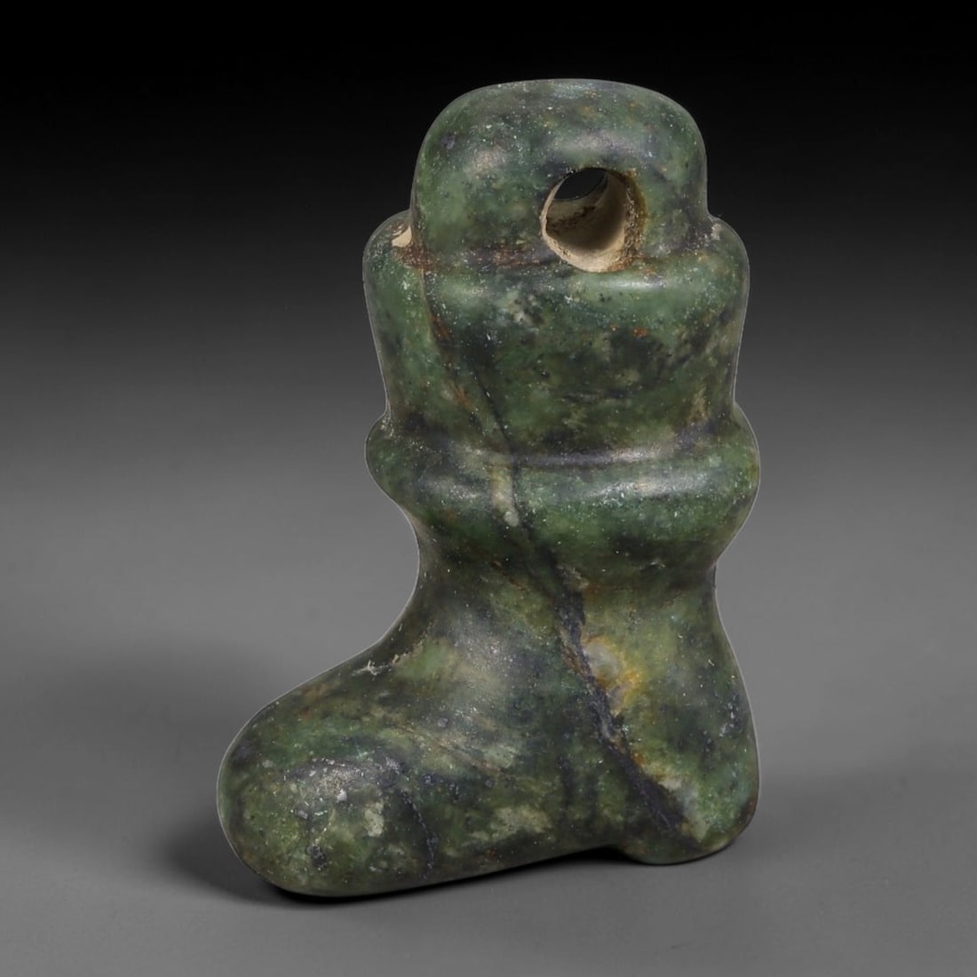 Ancient Western Asiatic Foot Form Ritual Amulet (1000-600 BCE), 93AE: Hand-carved mottled dark green serpentine pendant shaped as a stylized boot/foot motif with stacked rounded collars along the upper shaft. A large integrated pierced suspension loop at the top enables