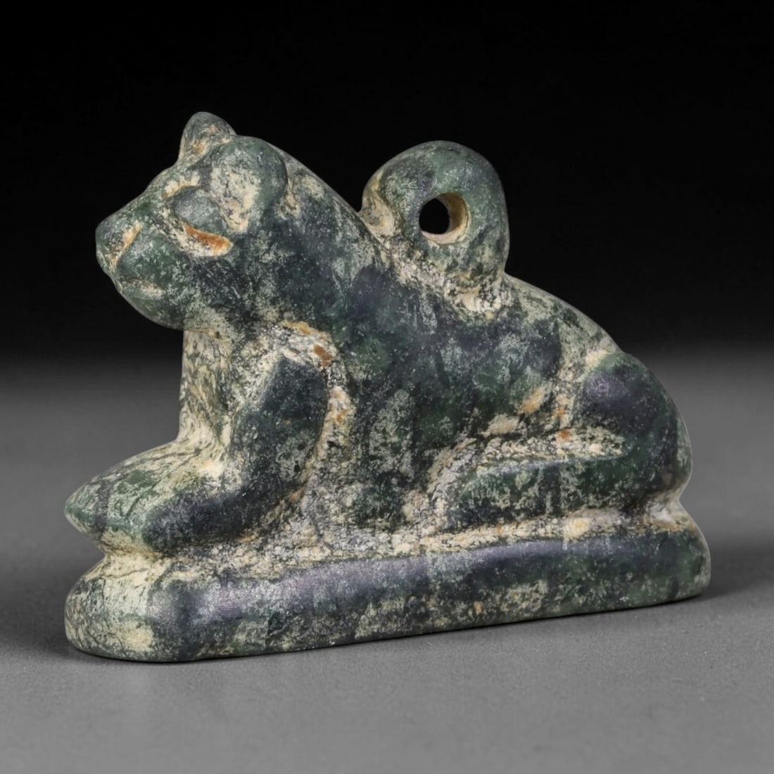 Ancient Egyptian Dark Green Serpentine Recumbent Feline Protective Amulet (664-30 BCE), 16AB: Hand-carved mottled dark green serpentine talisman depicting a resting lioness feline atop a flat integrated rectangular base. A large pierced suspension loop rises from the rear torso for cord wear; 